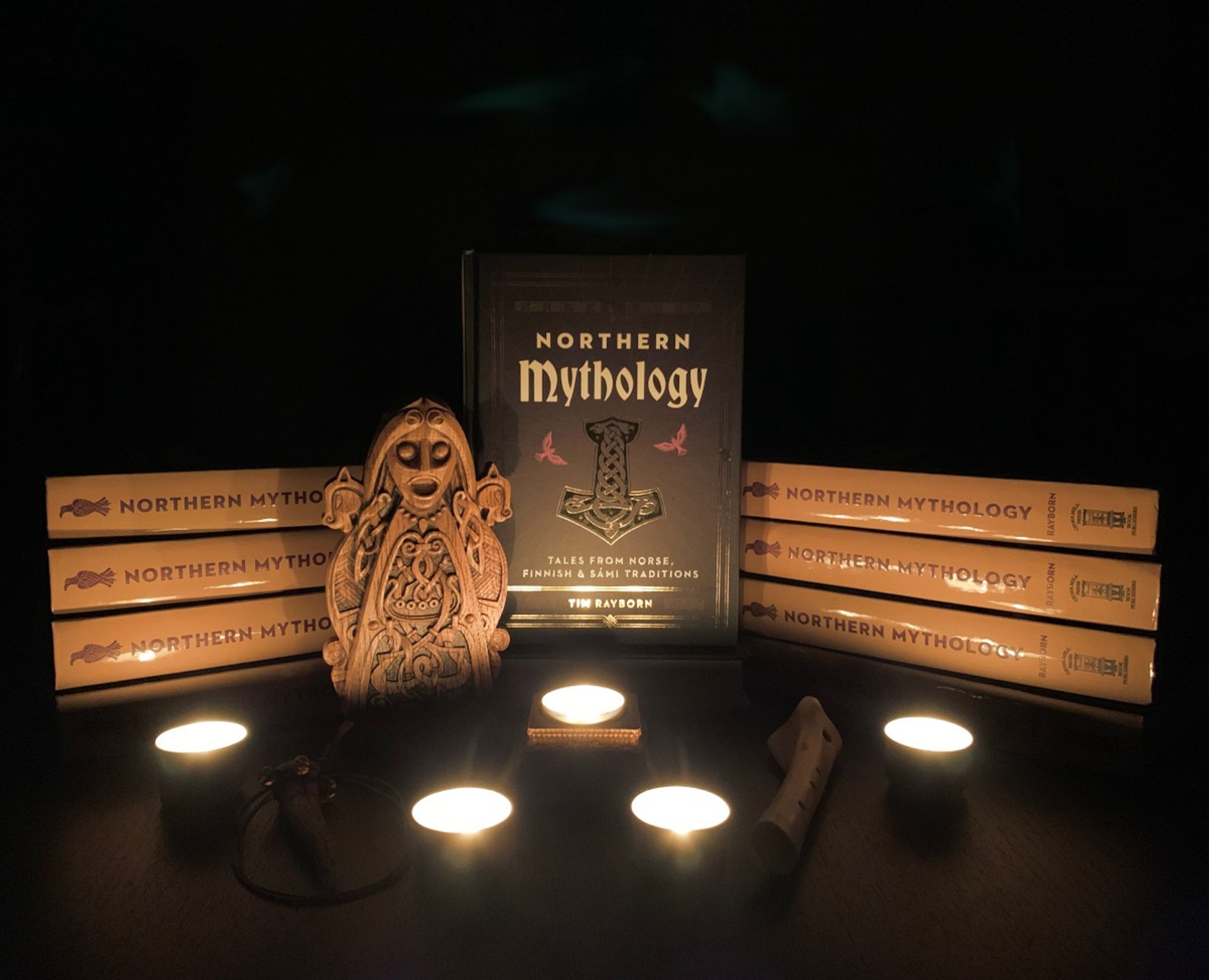 An image of copies of a book titled Northern Mythology are stacked in darkness, lit by candles. The image also features a beautiful old Norse, wooden statue of the goddess Freya, lit up by warm candle glow.