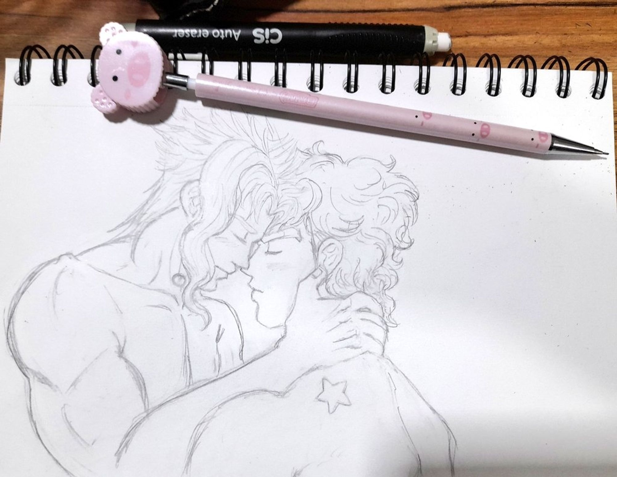 Pencil drawing of Jotaro and Kakyoin, they're naked, Kakyoin is holding Jotaro's neck and they're with their faces together, noses touching, very intimate. The drawing was made in a sketchbook and it's over a table. A mechanical pencil decorated with pigs and a eraser are on the top of the sketchbook.