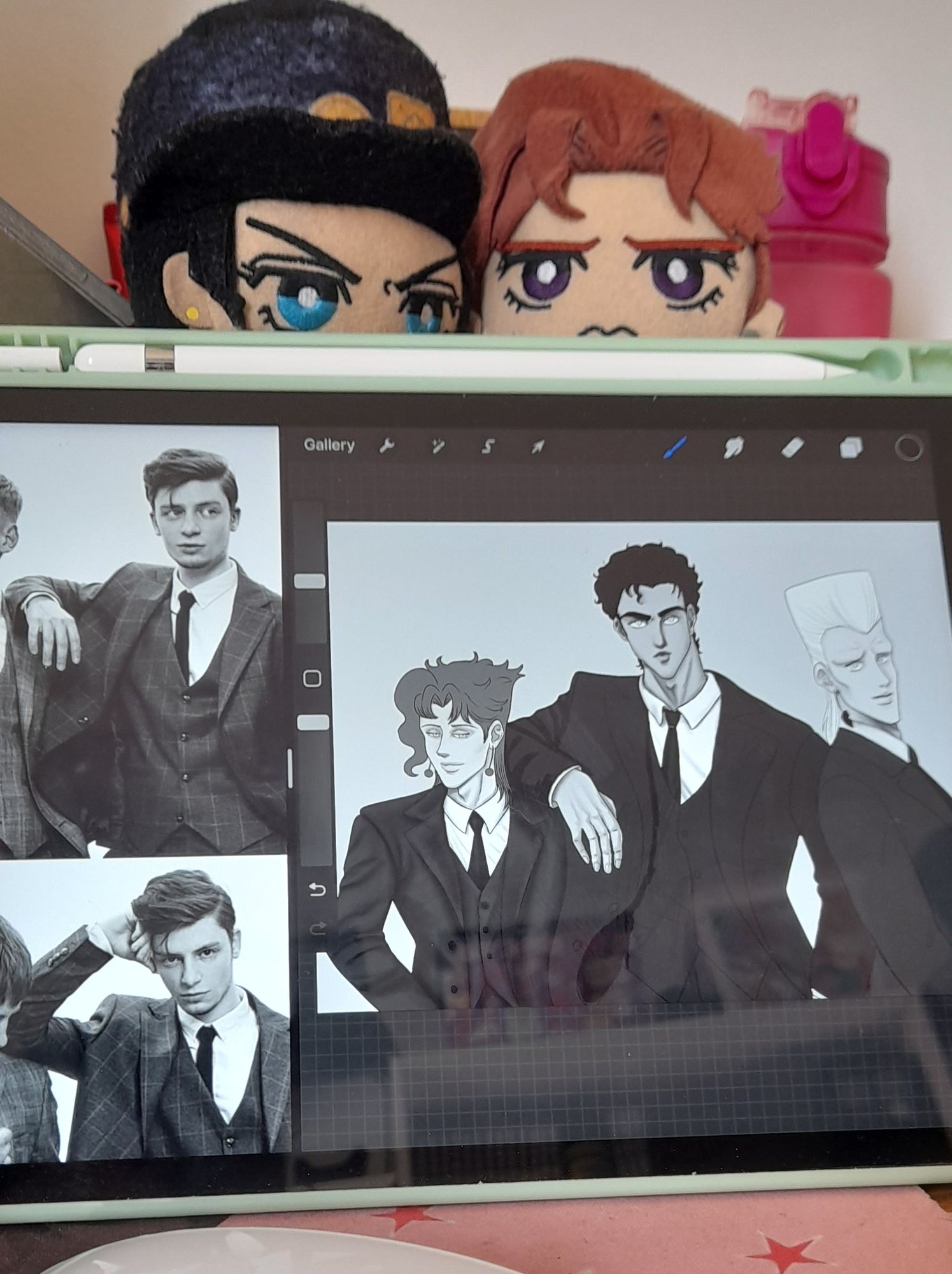 An ipad with a drawing of Kakyoin, Jotaro, and Polnareff wearing suits. The drawing is black and white and on the left side of the screen there's a photo that is the reference. On the background, behind the ipad it's possible to see Jotaro and Kakyoin plushies and a water bottle.