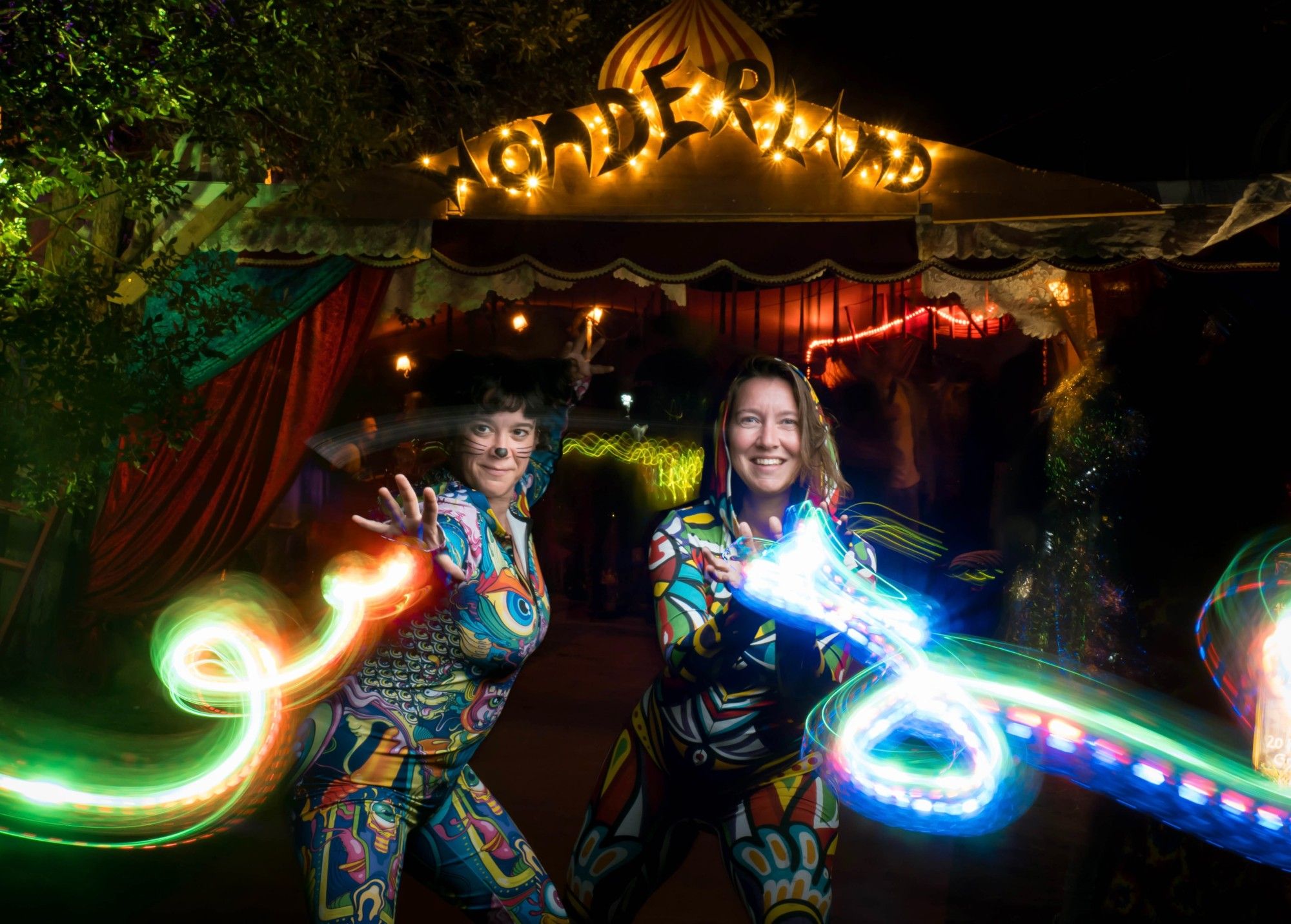 Lightpainting of two women in colorful clothes shooting lights from their hands. They stand in an archway with the word "Wonderland" written above.