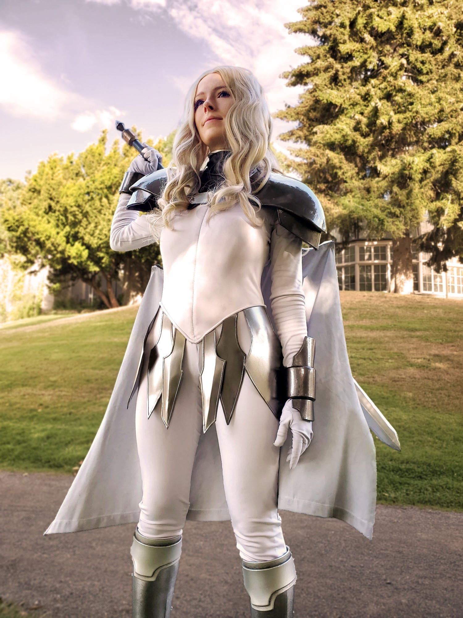 A woman standing triumphantly with one hand on the hilt of a broadsword sheathed at her back. She is cosplaying the character Teresa of the Faint Smile from the anime and manga series "Claymore". The sky is bright and the costume features glossy, dark steel piecemeal armor over the wrists, shoulders and waist, atop an off white body suit and cape.