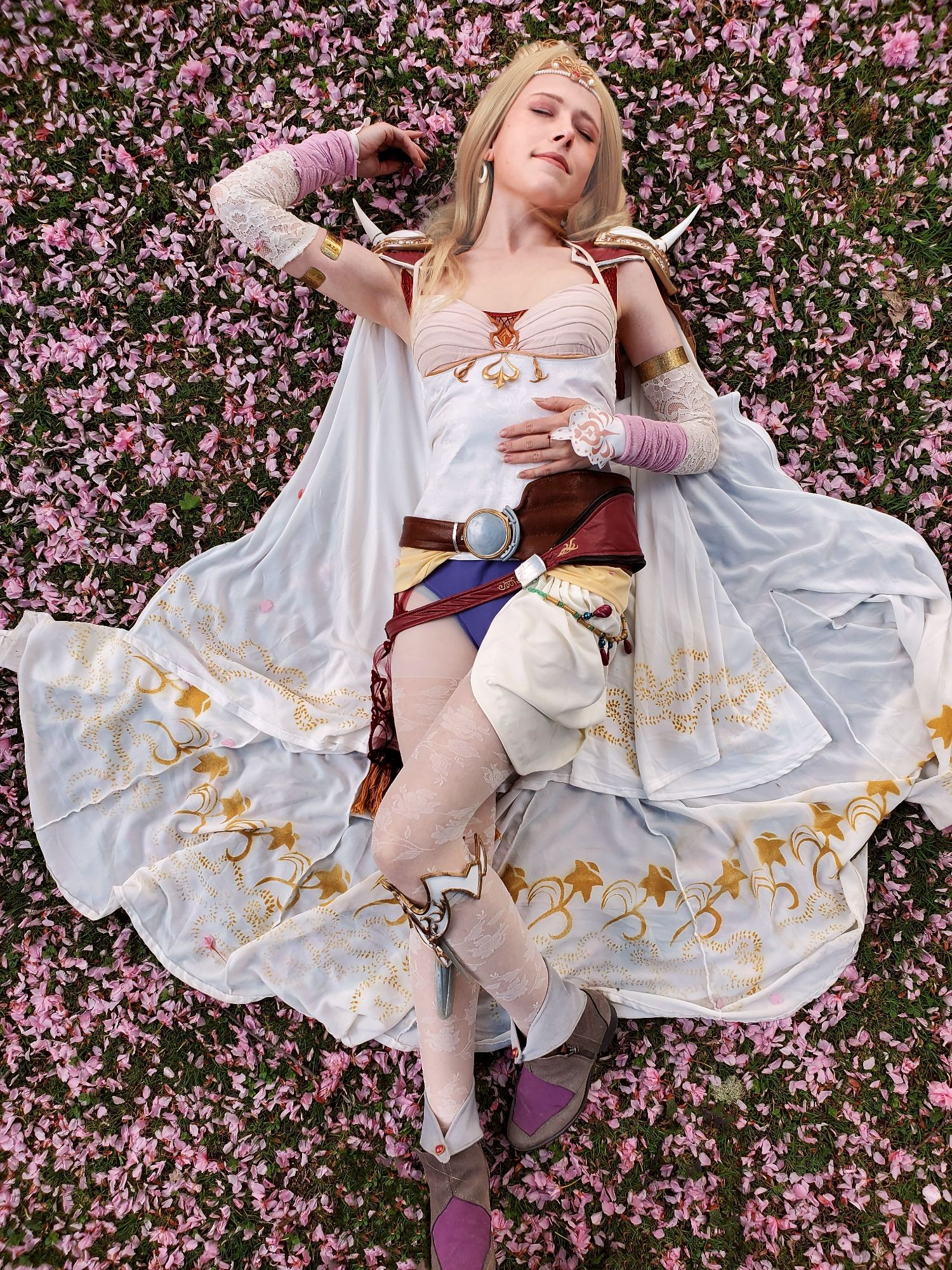 A woman in a complex patterned body suit lying elegantly in a field of pink Sakura flower petals. A white cape with gold floral accents fans out beneath her. The image over all has delicate pink, white and gold tones that bring out the romantic and almost royal feelings associated with the character she is cosplaying: Rosa Farrell from the video game Final Fantasy 4.