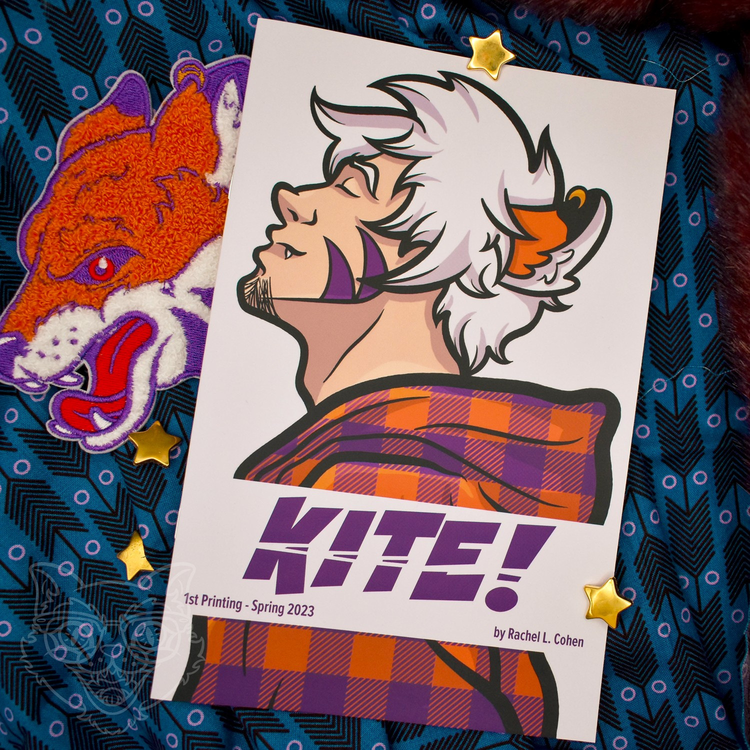 Cover of a zine on purple fabric featuring a side shot white haired young man with fox ears and an orange & purple plaid hoodie. Behind the zine is an embroidered patch of a fox head.