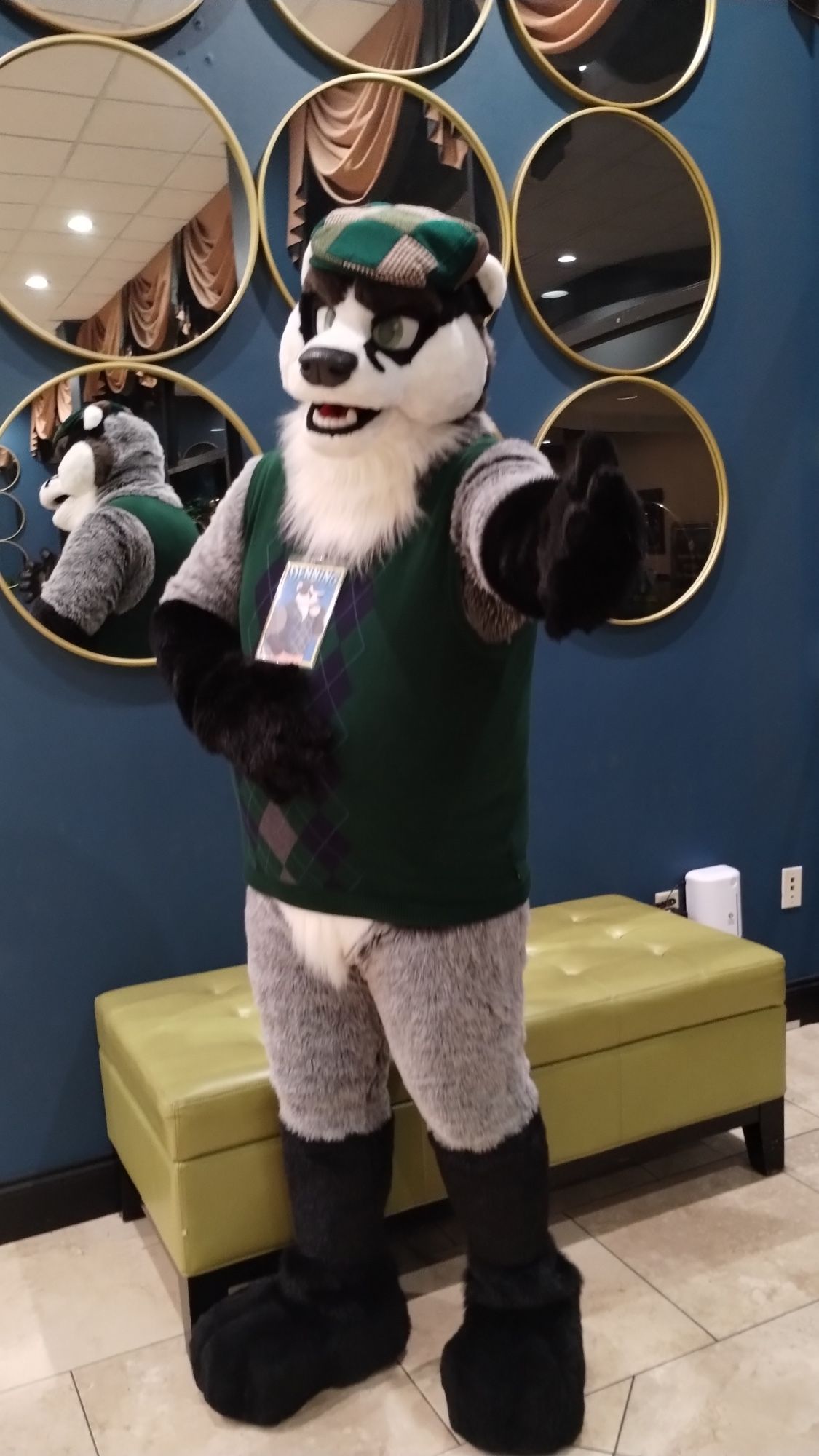 Badger fursuit, green flat cap and argyle sweater, in front of mirrors in hotel lobby, one hand on tummy and one hand outstretched