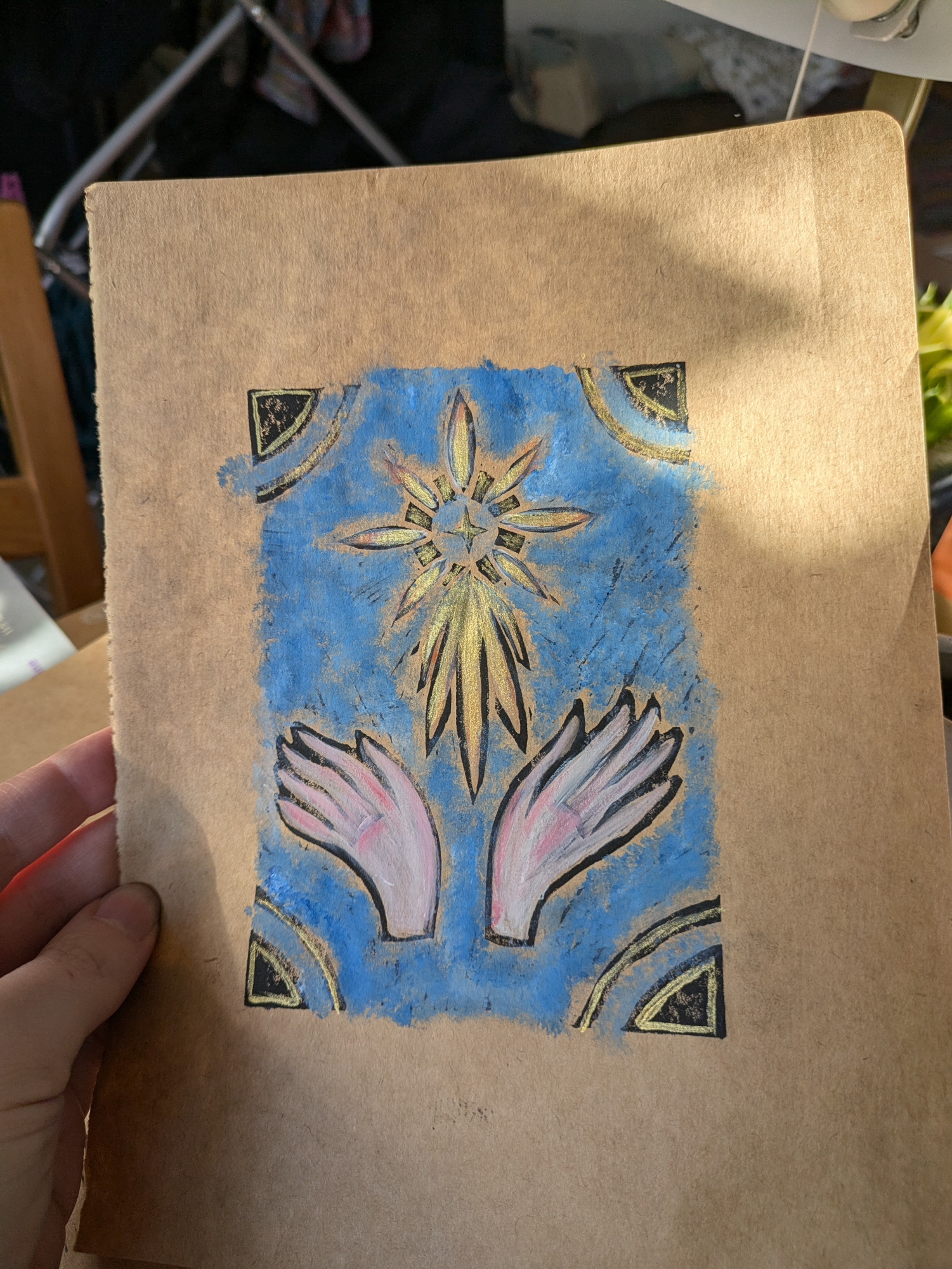 A photo of a linocut print of a pair of wings or hands pushing a shooting star upwards. It's printed on brown paper with black ink, with colours added in acrylic paint: white and pale pink for the wings, gold on the star, and a dappled blue background.