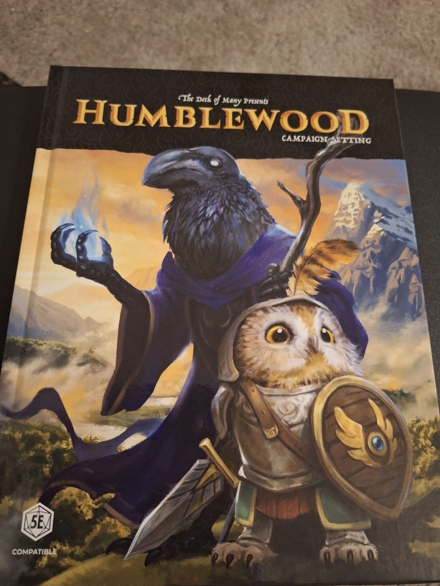 Photo of the Humblewood campaign setting hardback for 5e. There is a tall raven mage and a smol owl paladin in a mountainous setting. I am going to learn what they're actually called by reading this book.