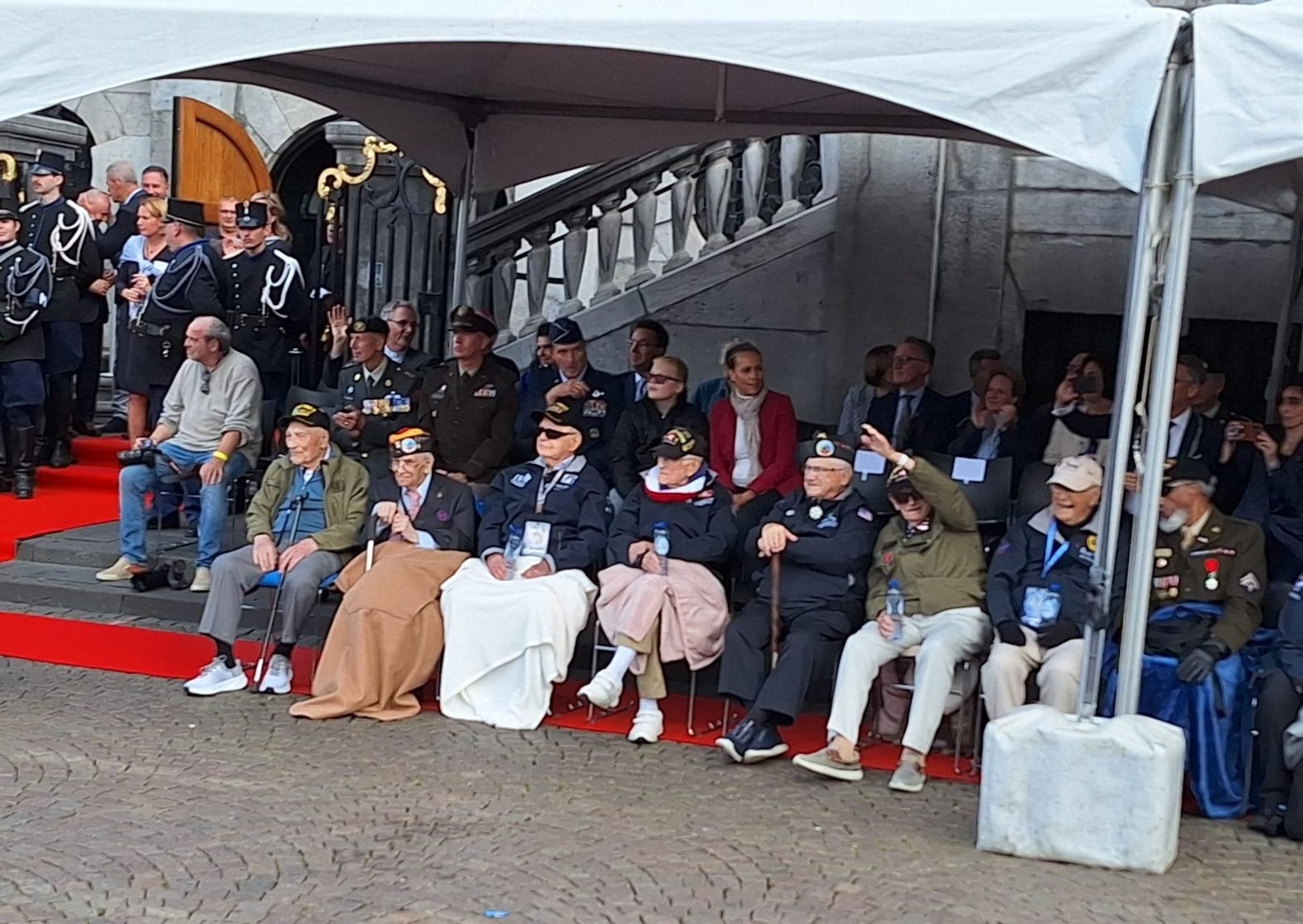 Acht veteranen, zittend op een rij, kijkend naar de Liberation Parade, zwaaiend naar deelnemers en publiek.