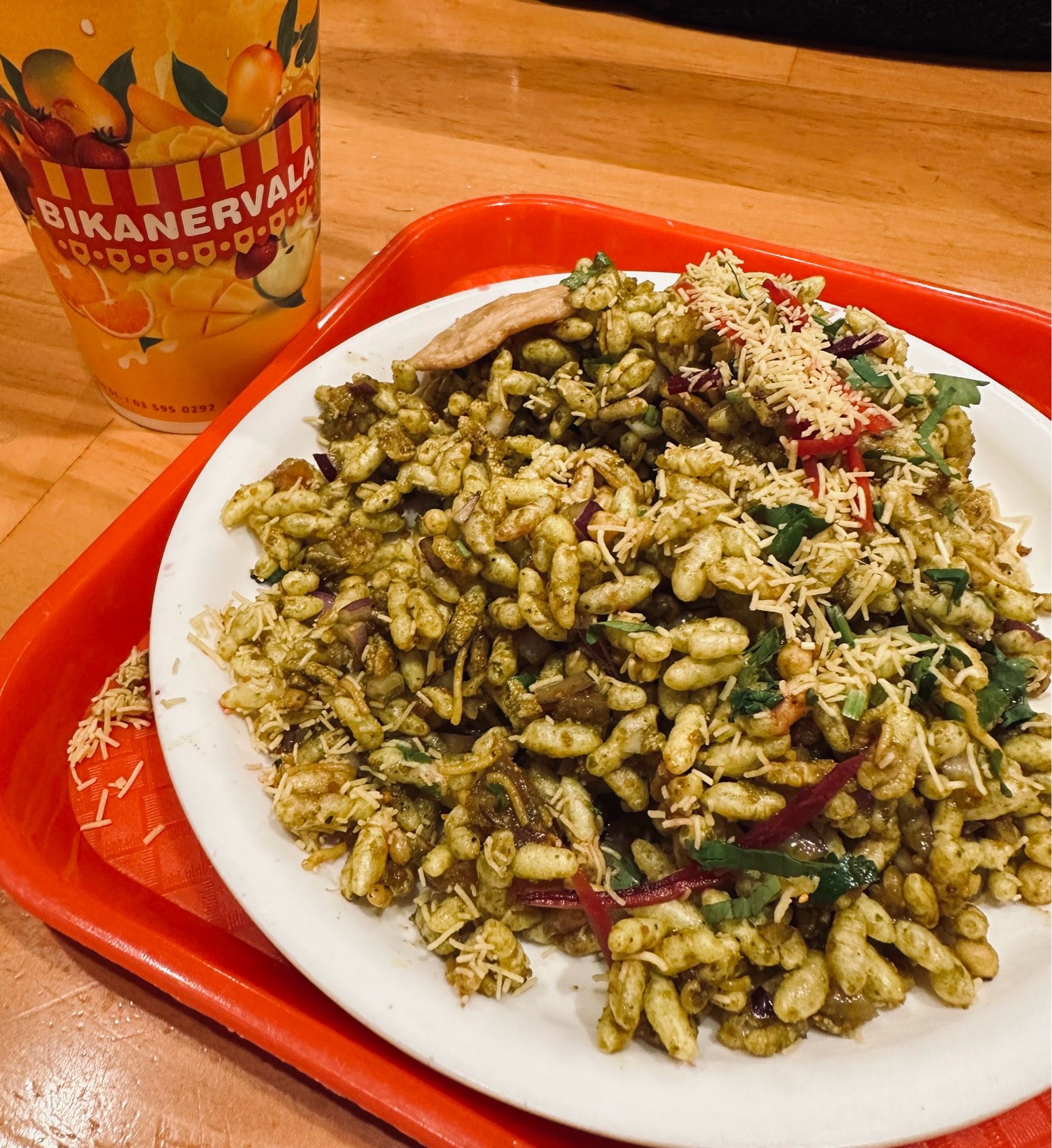 White plate of Indian Bombay Bhel Puri (puffed rice savoury food) on a red tray. Cup of mango lassi on left side branded in bright orange and red, ‘Bikanervala’.