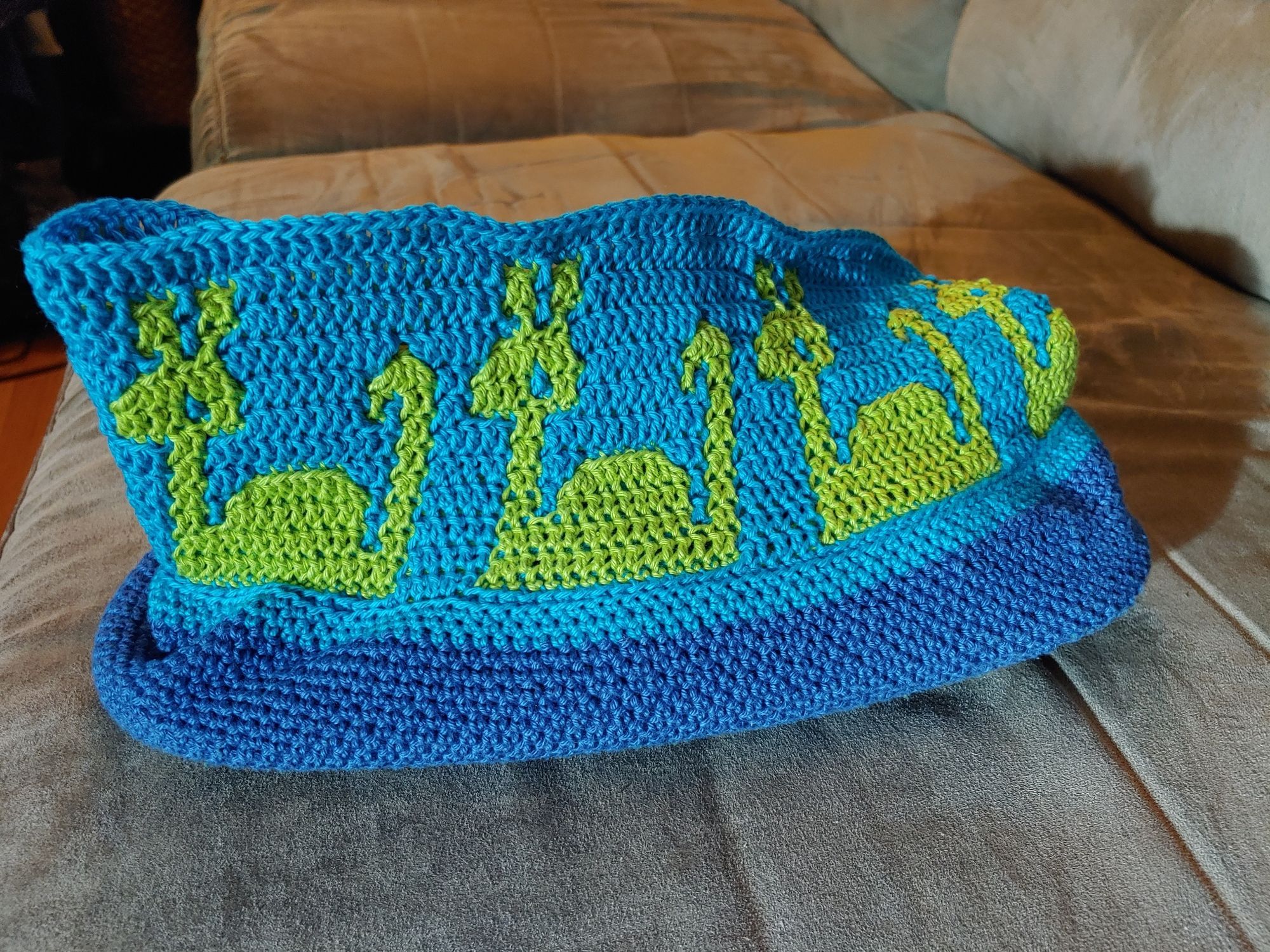 A work-in-progress crochet purse. The bottom is royal blue, with a teal blue & lime green repeating motif of a cute Nessie sea monster. She's in the green & the background is the teal blue. 4 Nessie motifs are visible on this side view.