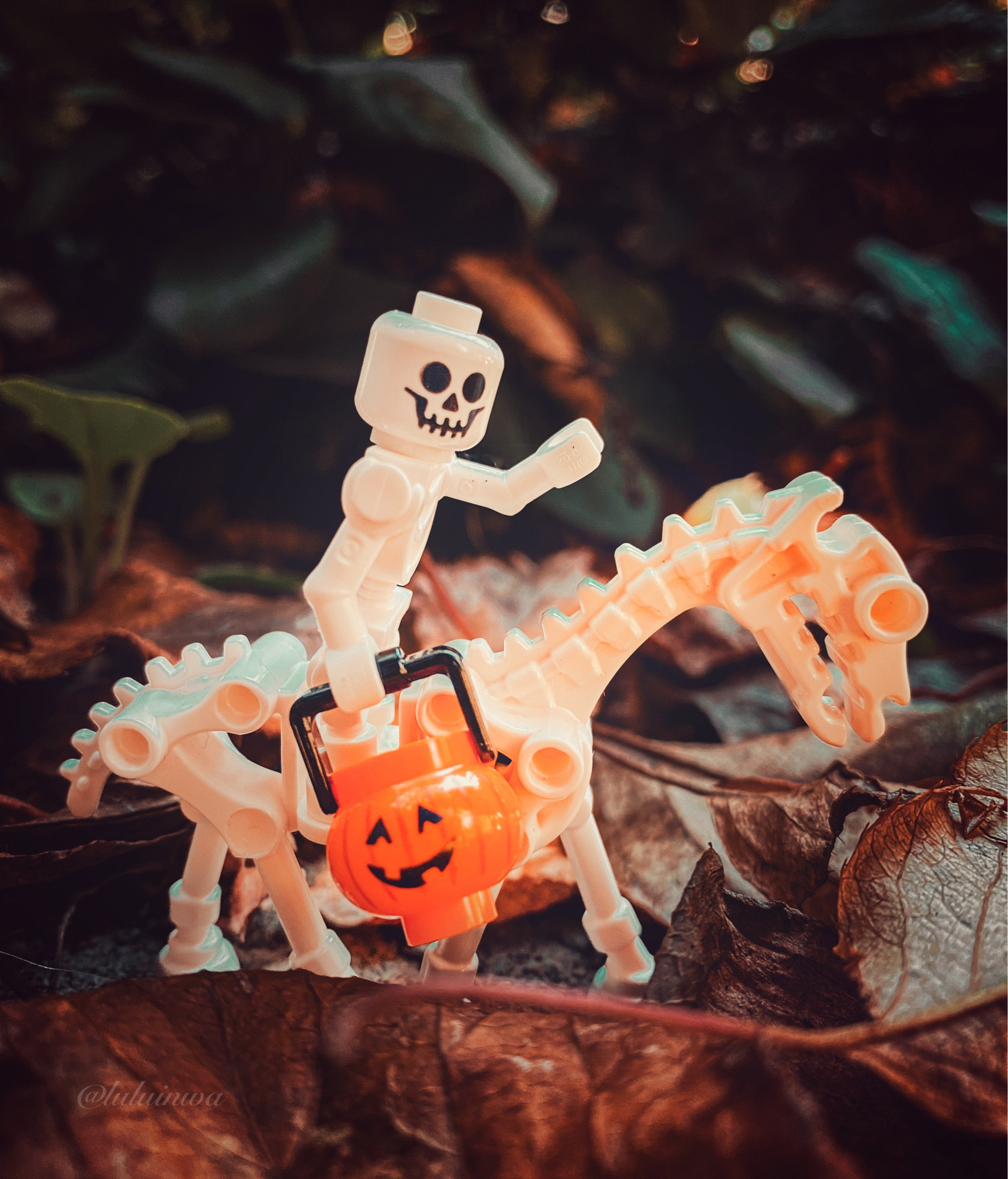 Lego skeleton holding a Jack-o-lantern riding a skeletal horse in a forest surrounded by fallen leaves.