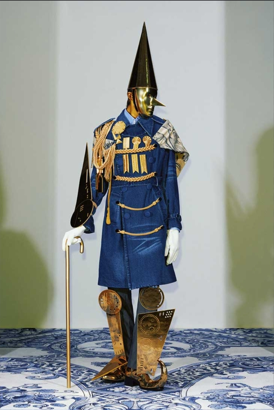 A model from the 2025 spring xander zhou collection. An exaggerated blue military style jacket with gold trim. Gold cutouts attached to legs, and gold cane