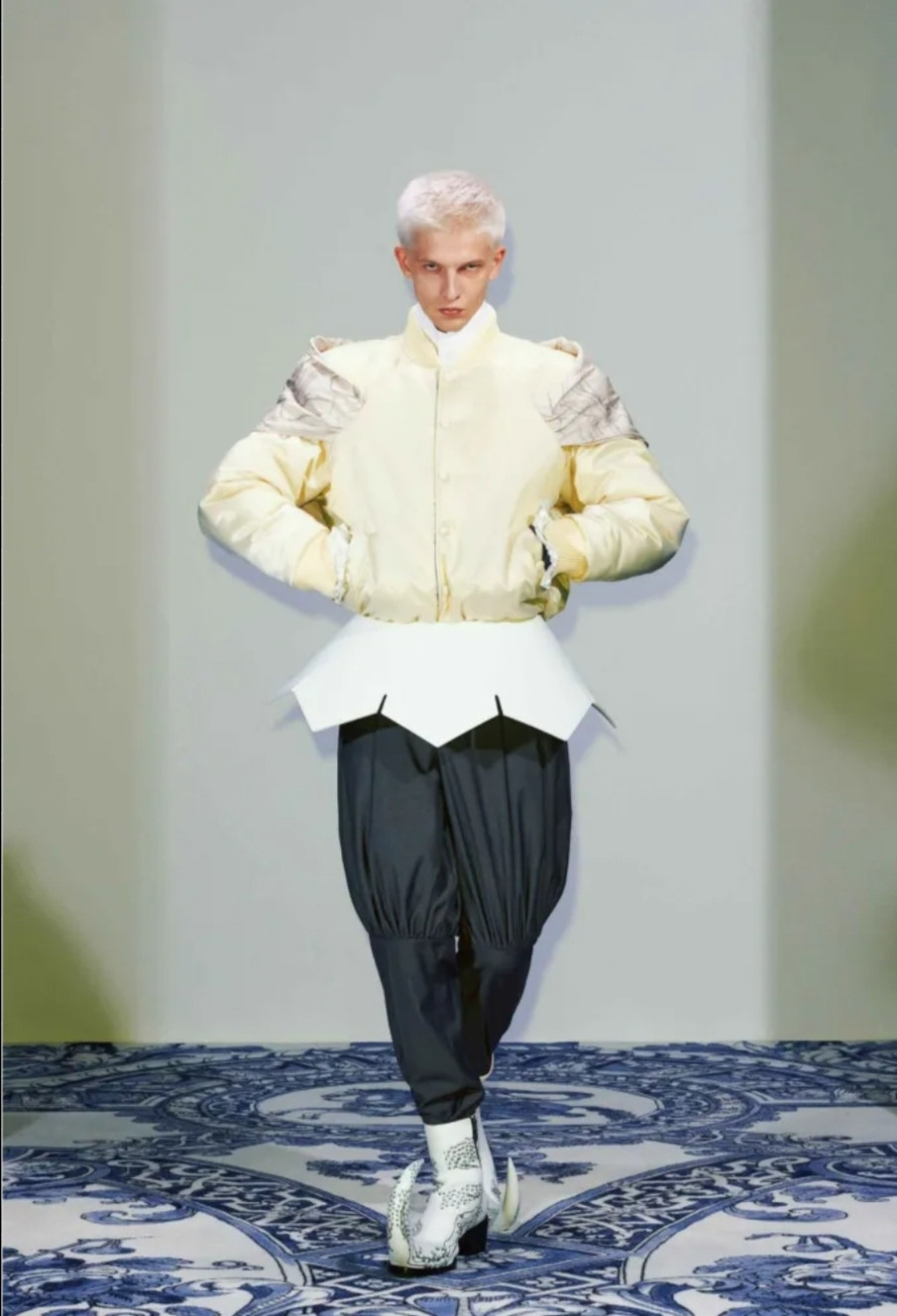 A model from the 2025 spring xander zhou collection. A white puffy jacket, gray trousers tailored to look like pantaloons. Silver pointy toed boots