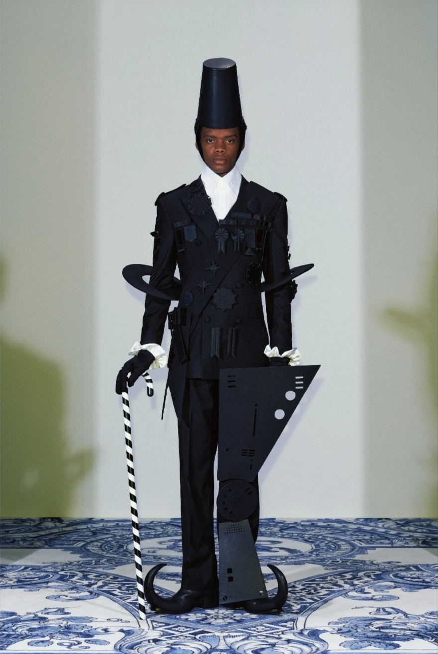 A model from the 2025 spring xander zhou collection. A black suit, white scarf and cuffs, black pointy toed boots, candy striped cane