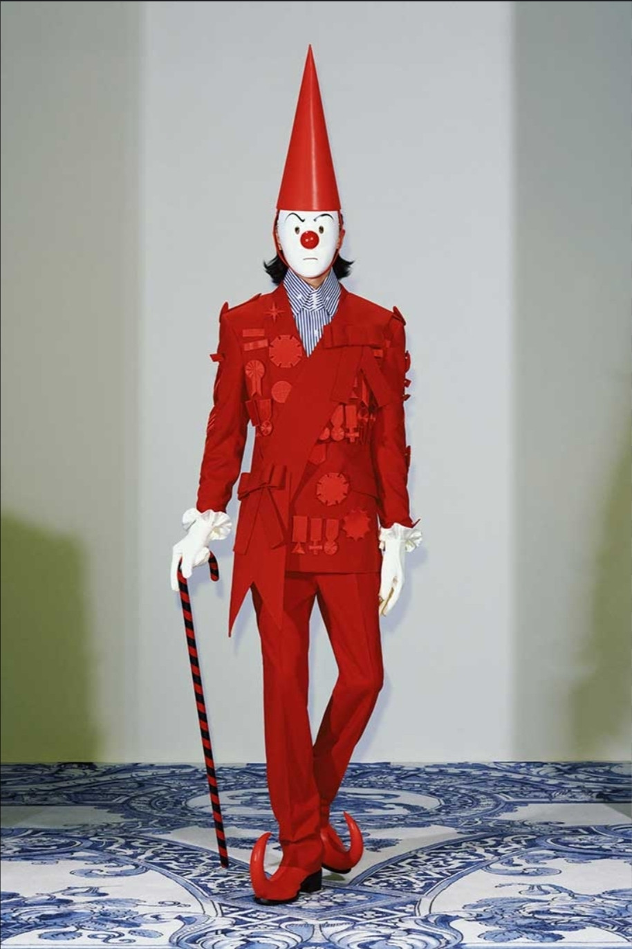 A model from the 2025 spring xander zhou collection. A red monochrome suit with pointy red boots, white gloves, clown mask and candy striped cane