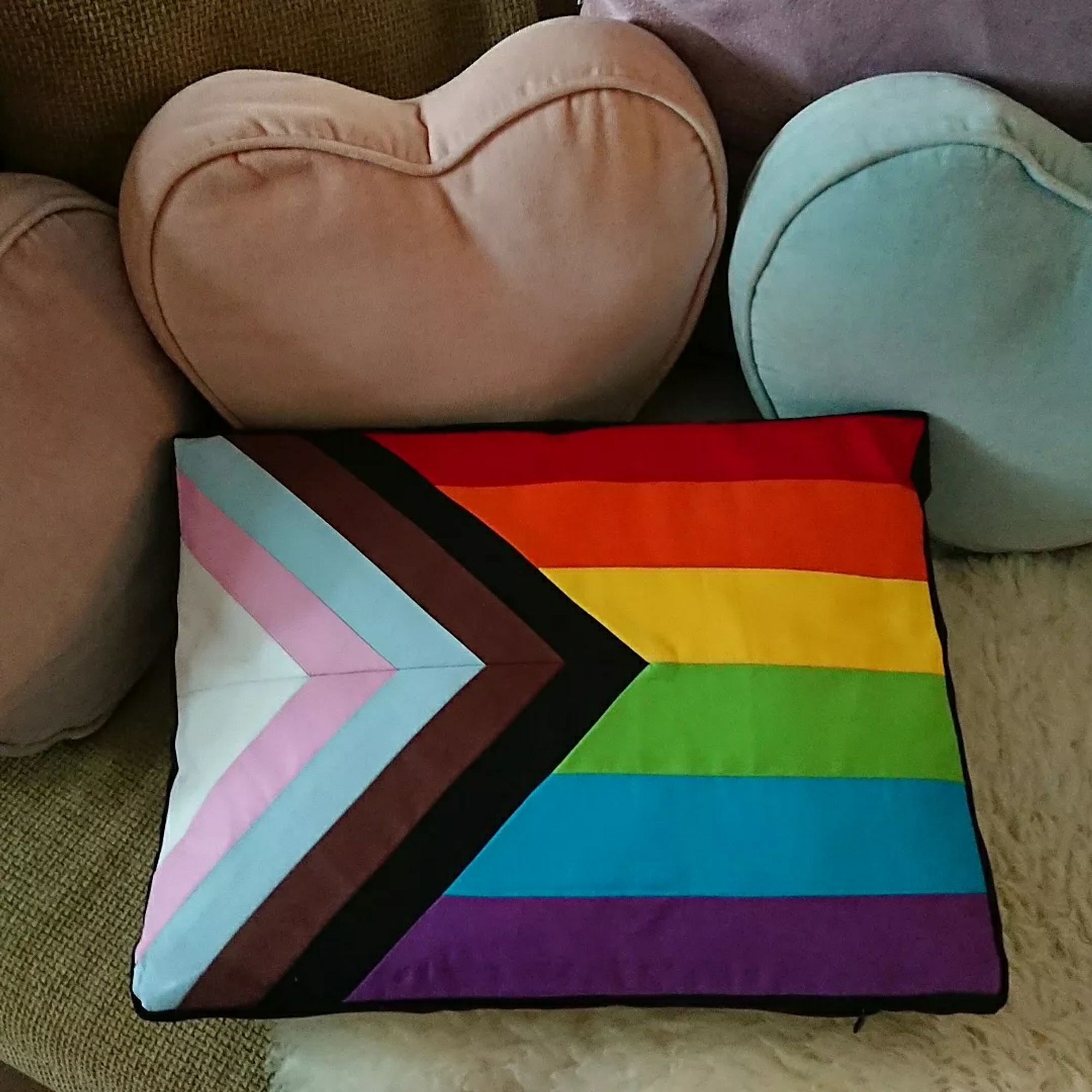 A rectangular shaped cushion with the Progress Pride flag on it. There are horizontal stripes in red, orange, yellow, green, blue and purple with an additional herringbone pattern along the right side featuring black, brown, light blue, pink and white stripes. It's leaning against three heart shaped cushions in purple, pink and blue, all on an olive green sofa with a cream sheepskin rug.