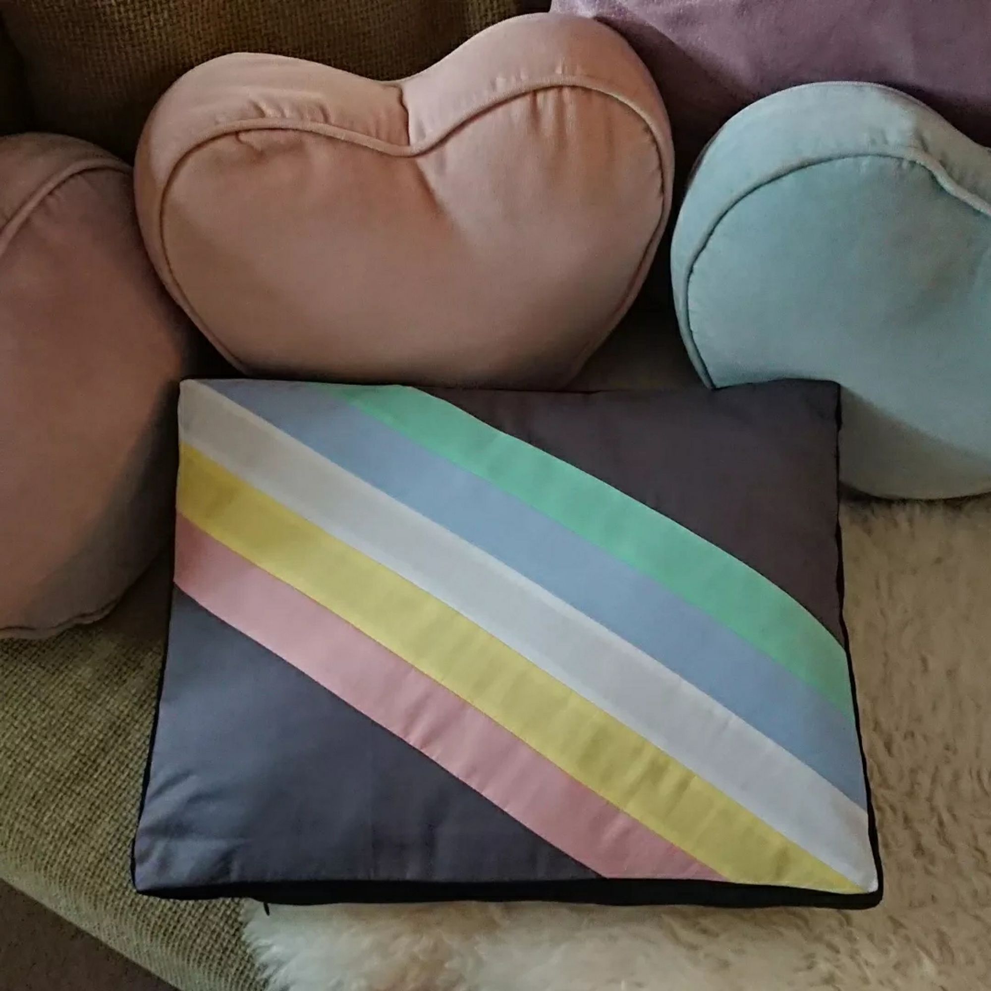 The other side of the rectangular shaped cushion on the same sofa, this side has the Disability Pride flag on it. It has a charcoal grey background with a diagonal band from the top left to bottom right corner, made up of five parallel stripes in softer tones of green, blue, white, yellow and red.