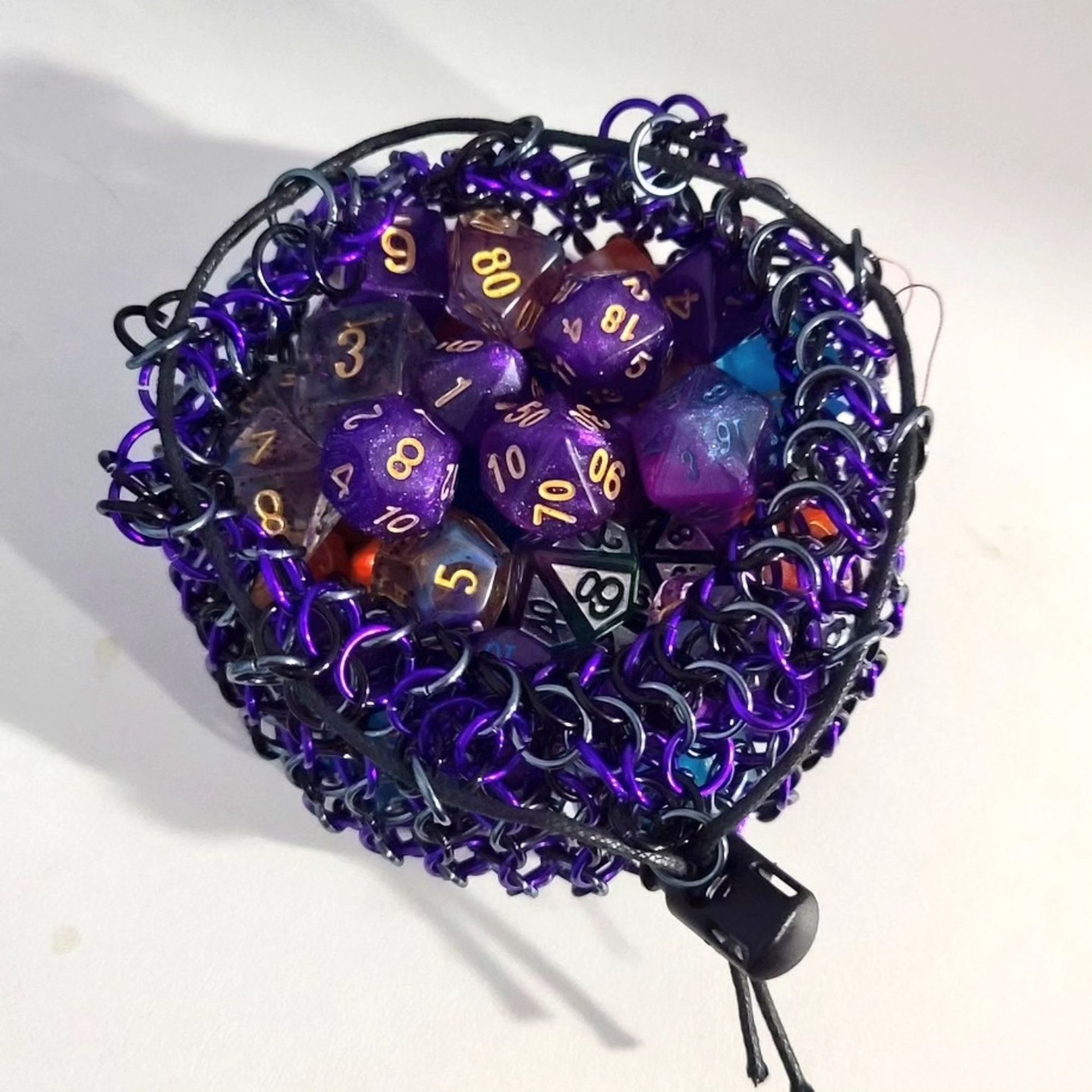 a picture of a chainmail dice bag made of black, purple, and gray rings. It has a black cord and toggle clasp for closure. It is open and full of dice. the bag is resting on a white tabletop.