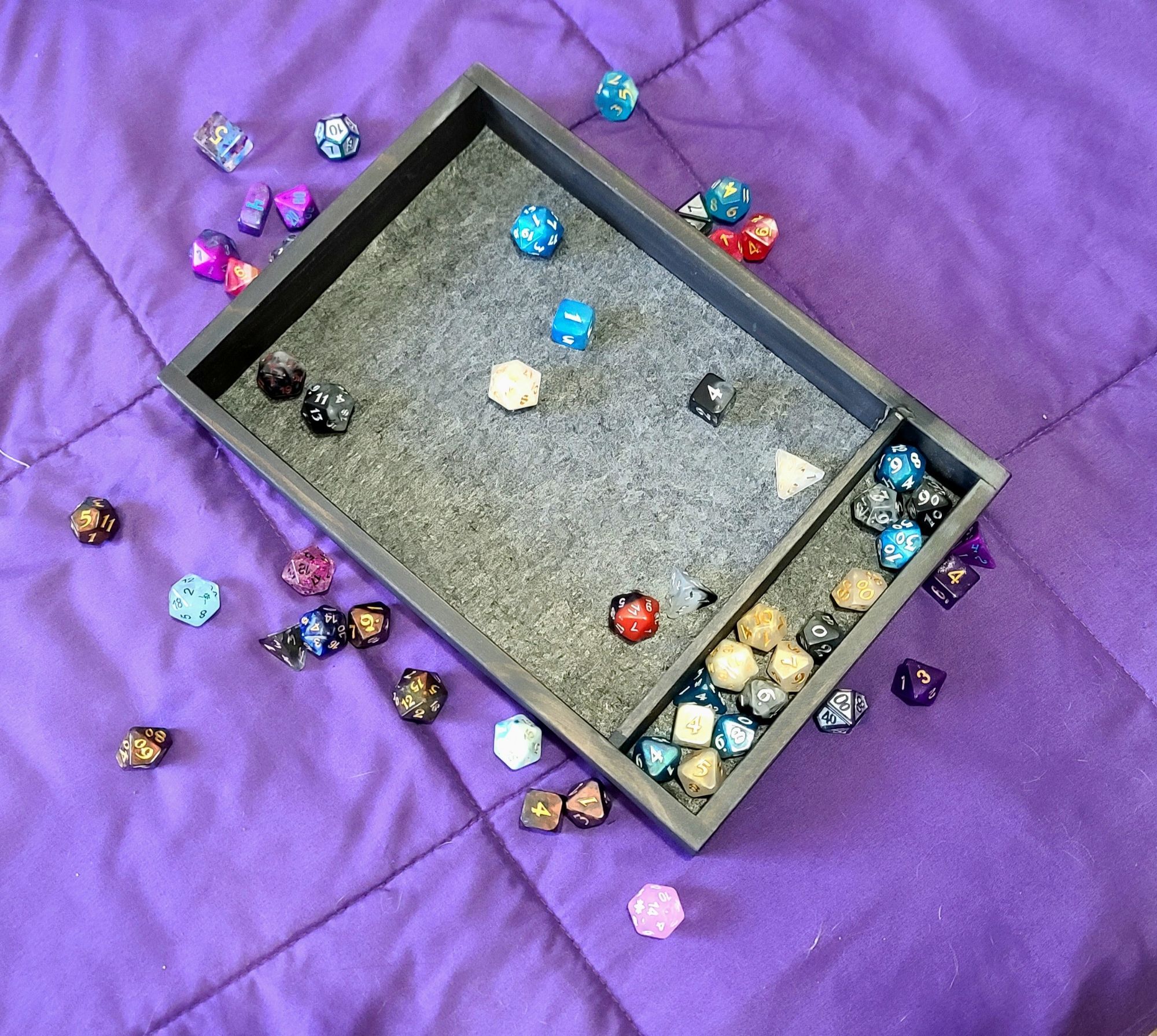 a picture of a wood dice tray painted with a black wash. it has a gray felt rolling surface. the tray is filled with dice and has dice scattered around the outside of it. it is resting on a purple blanket.