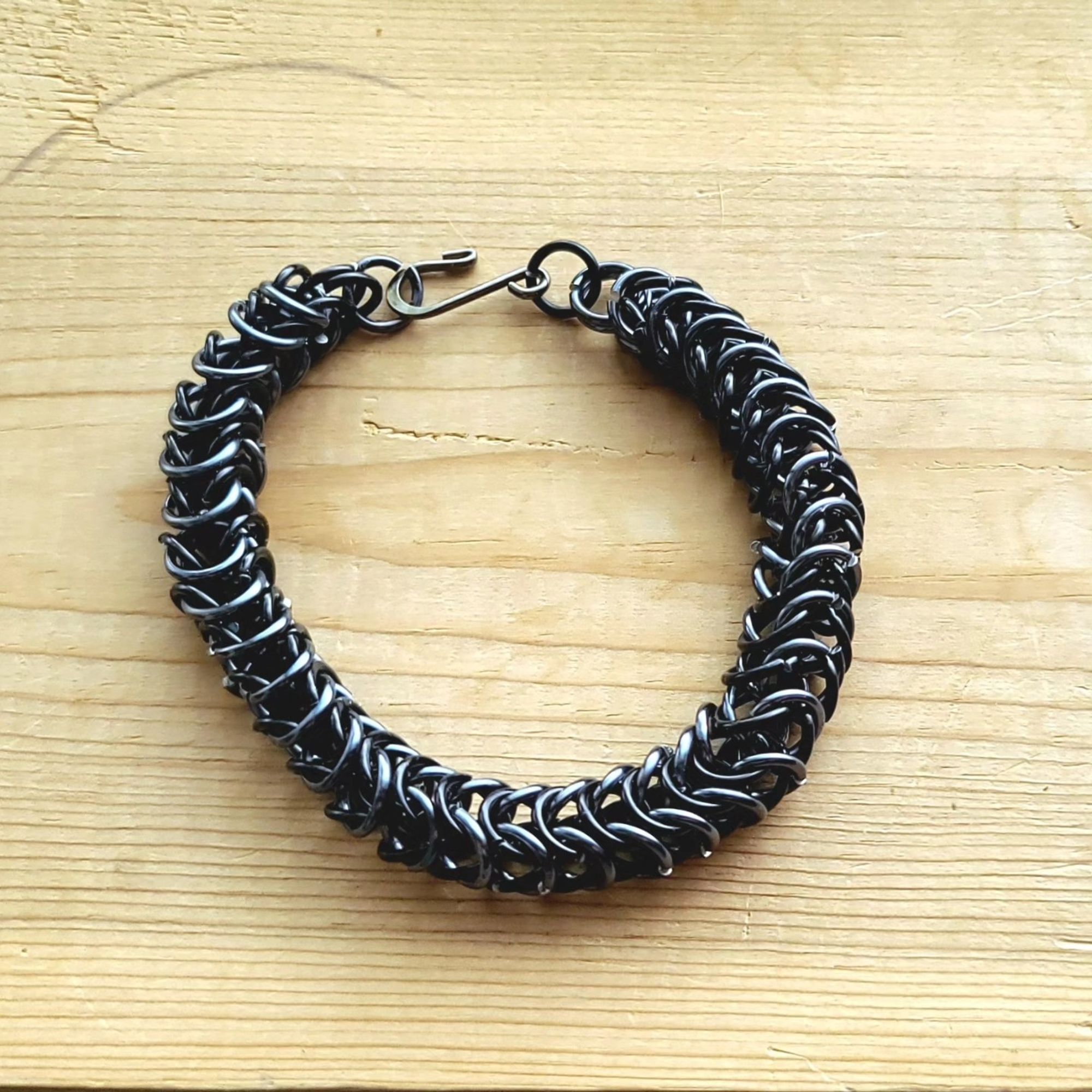 a picture of a box weave chainmail bracelet made with black and gray rings in an alternating pattern. it has a hook clasp. it is resting on a wood tabletop.