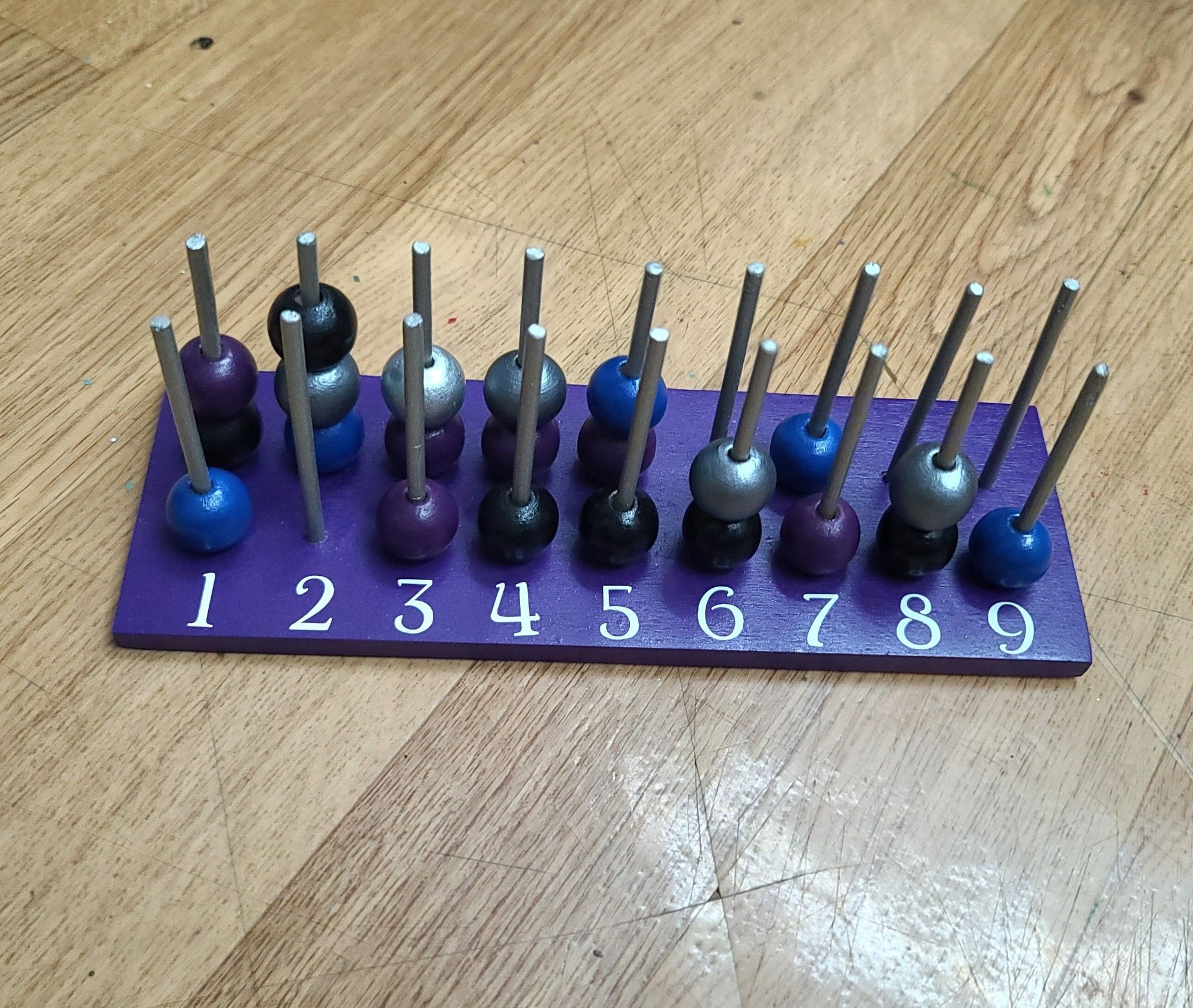 a picture of a wooden spell slot tracker. a rectangular piece of wood with 18 wooden pegs lined up in two rows of 9. it has several multicolored beads on the pegs and numbers 1-9 in front of the pegs. colors: purple bottom; silver pegs; white numbers; and silver, black, purple, and blue beads.