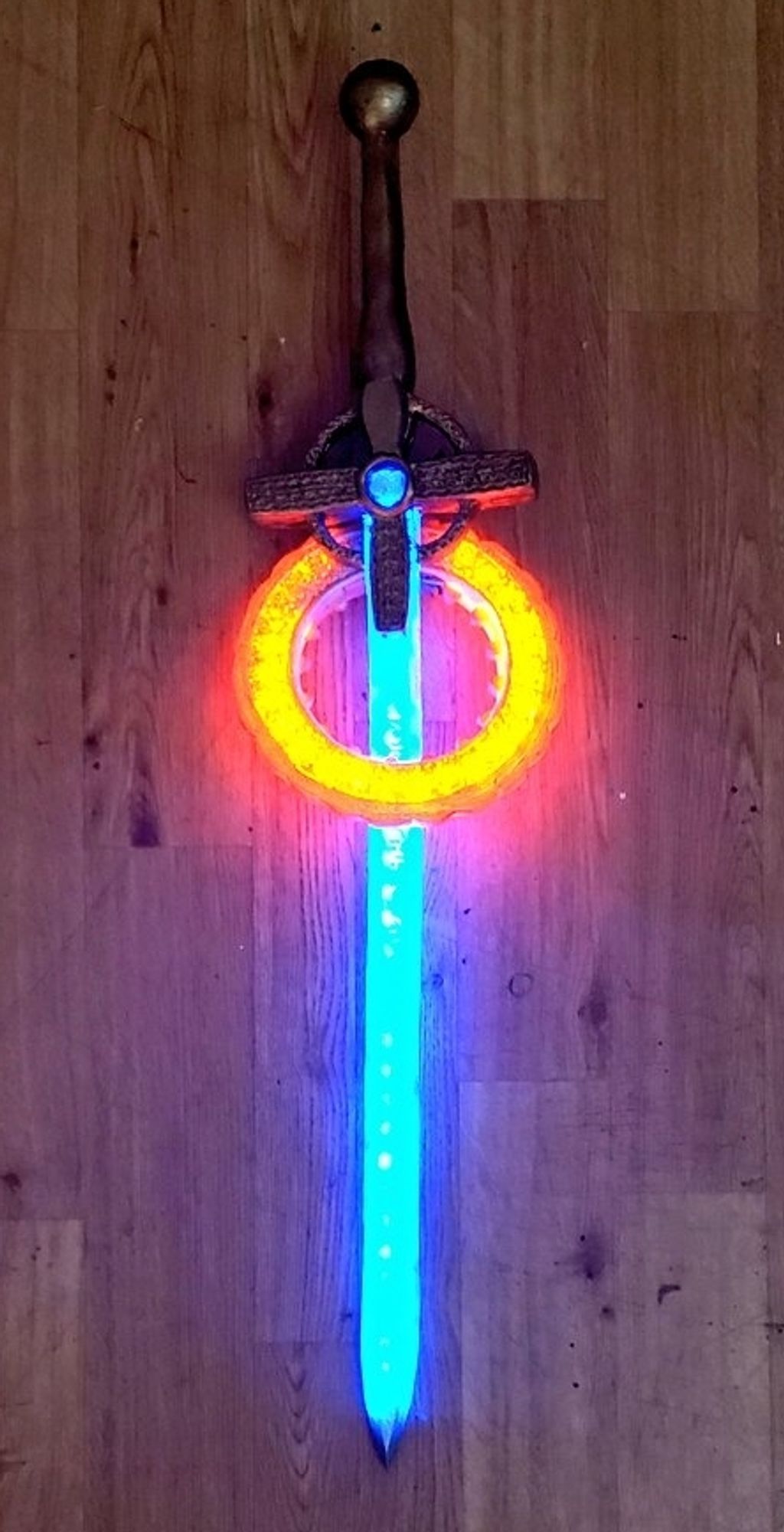 a foam prop replica of the cruciform sword and halo from the netflix series warrior nun. the room lights are off and the blade of the sword is lighting up blue and the halo is lighting up orange.