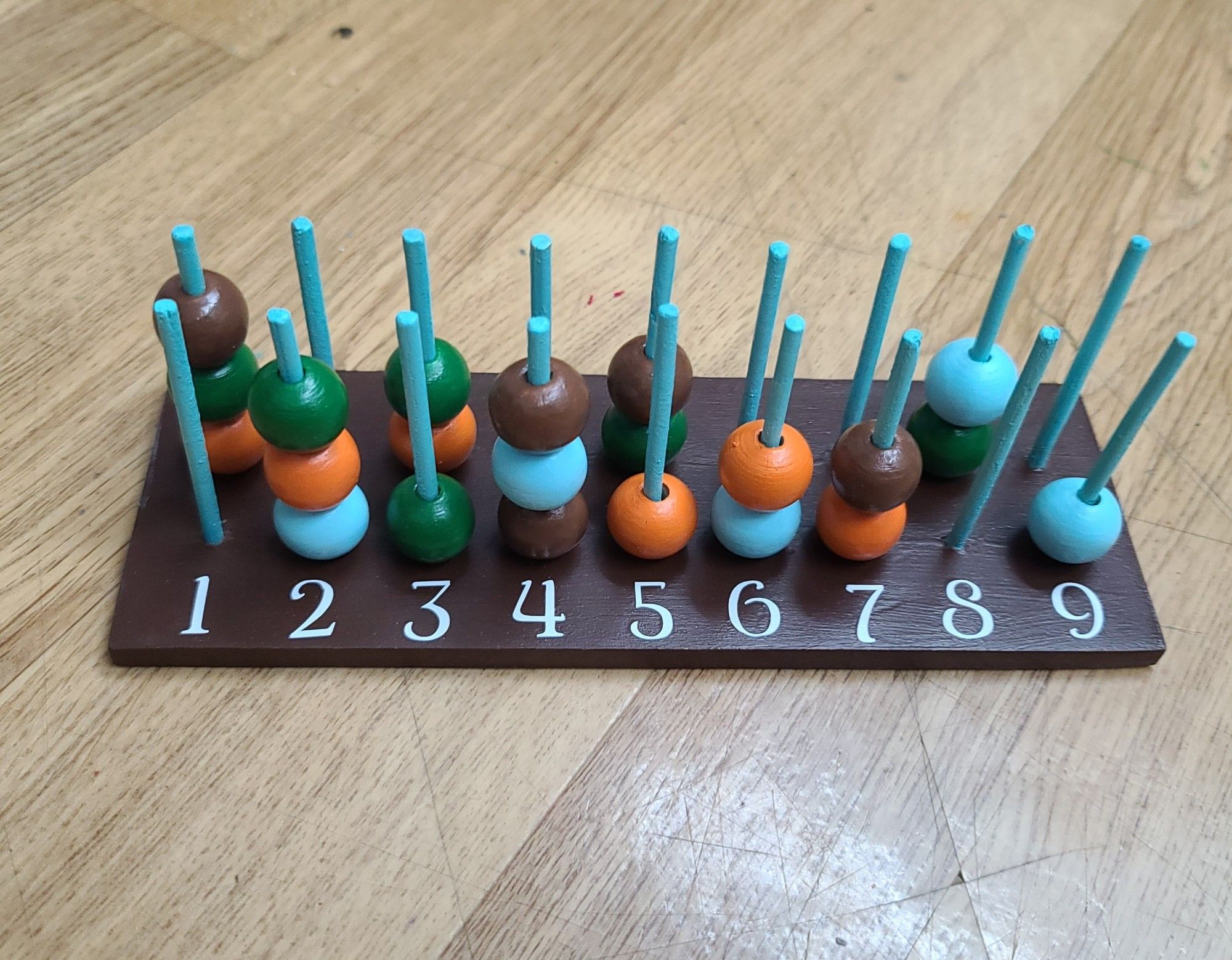 a picture of a wooden spell slot tracker. a rectangular piece of wood with 18 wooden pegs lined up in two rows of 9. it has several multicolored beads on the pegs and numbers 1-9 in front of the pegs. colors: brown bottom; light pegs; white numbers; and light blue, brown, orange, and green beads.