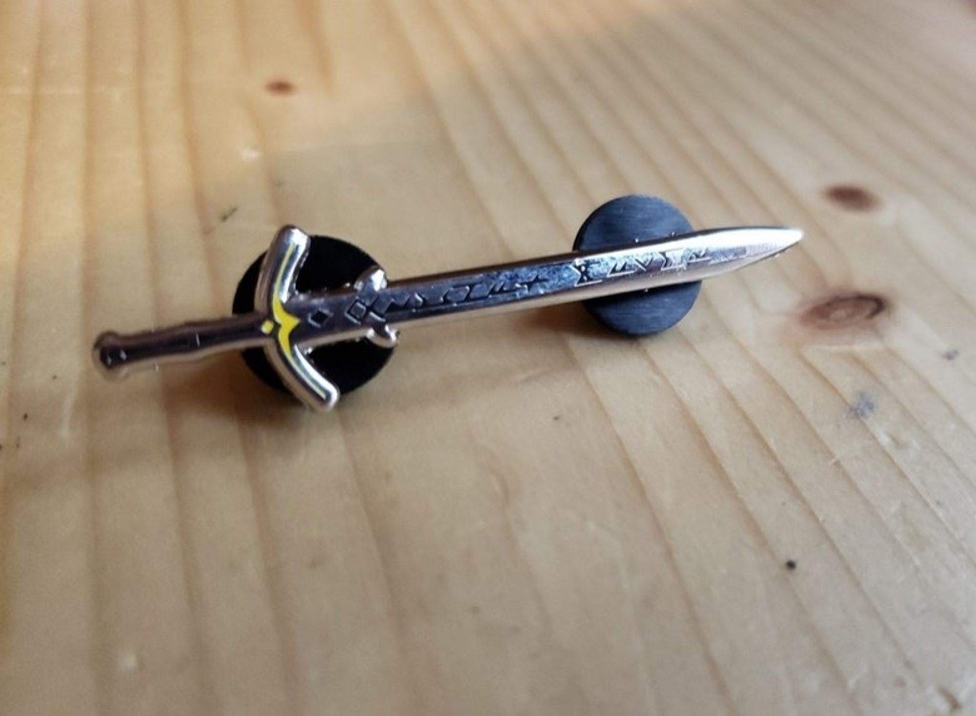 a picture of a sword enamel pin. the sword design is a zweihander greatsword with runes spelling out "orphan maker" down the blade. the pin js resting on a wood tabletop.