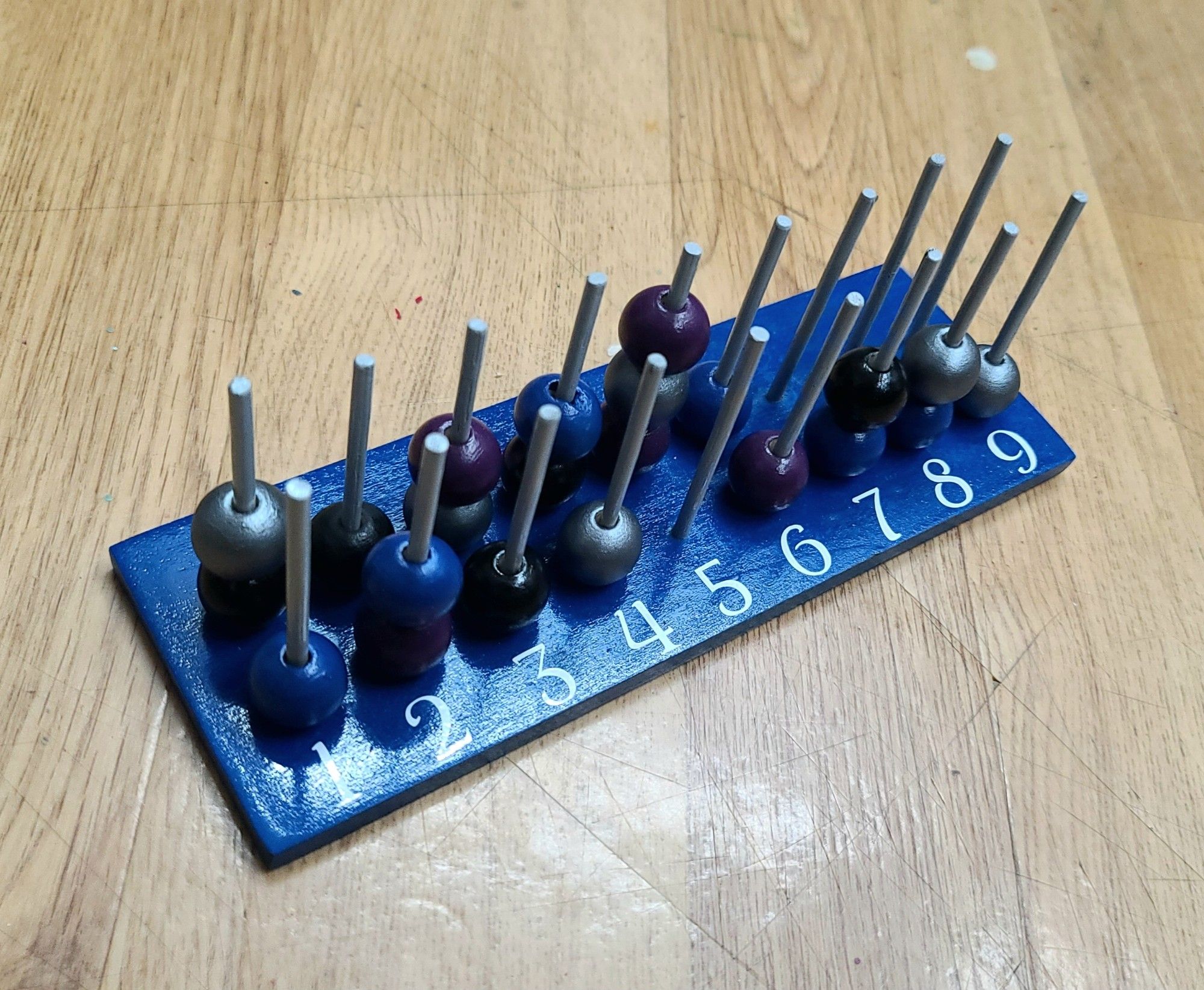 a picture of a wooden spell slot tracker. a rectangular piece of wood with 18 wooden pegs lined up in two rows of 9. it has several multicolored beads on the pegs and numbers 1-9 in front of the pegs. colors: blue bottom; light gray pegs; white numbers; and silver, black, purple, and blue beads.