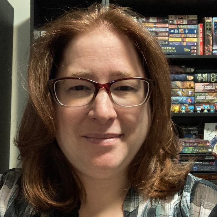 woman with red hair cut just past the shoulders wearing glasses in front of a bookshelf.