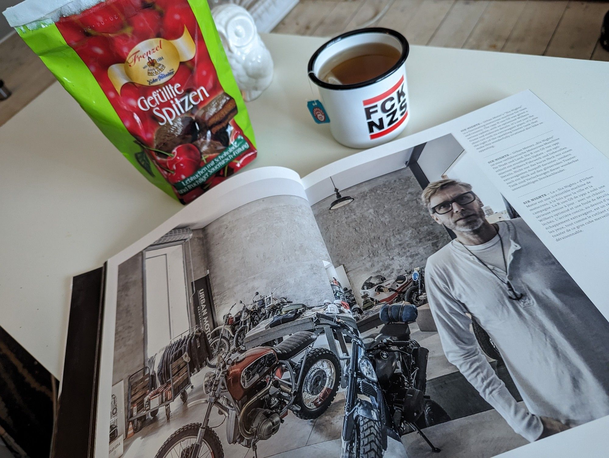 Ein Couchtisch auf dem ein aufgeschlagenes Buch über Custombikes liegt. Daneben eine volle Teetasse mit FCK NZS Logo und eine Packung gefüllte Lebkuchenspitzen