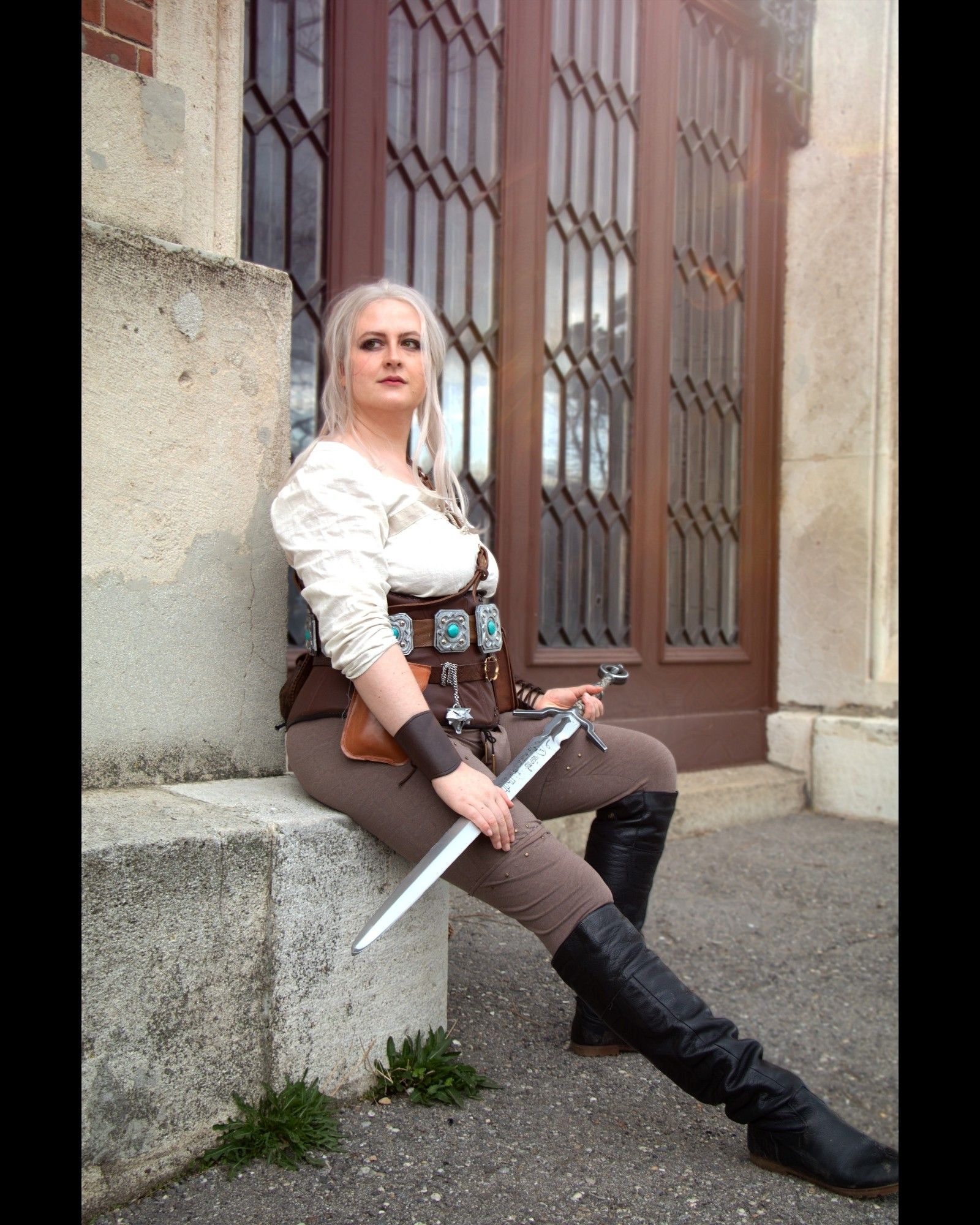 Me in cosplay as Ciri sitting against a wall, holding her sword on her lap and looking wistfully off into the distance