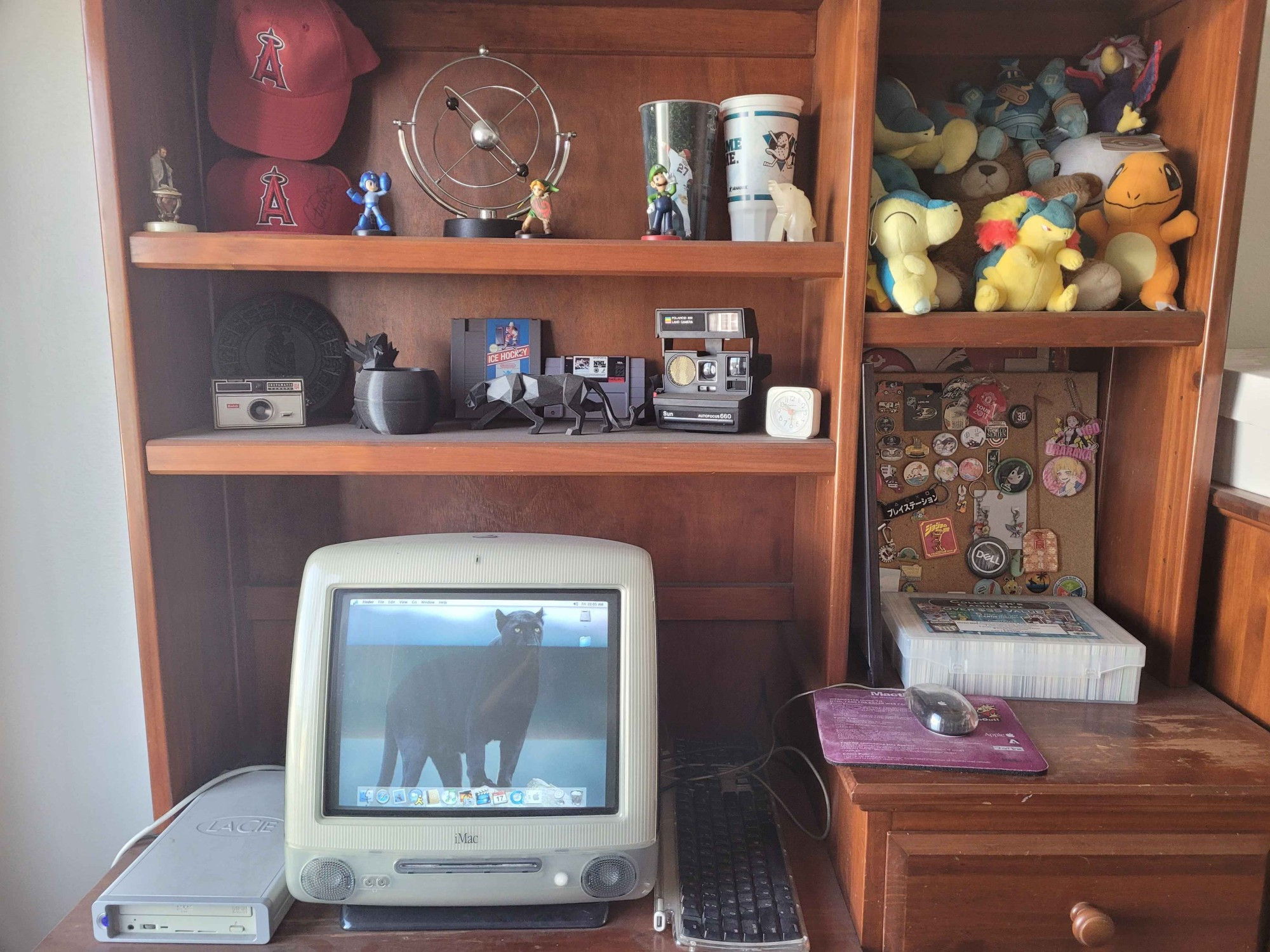 A picture of a corner of my room that shows off many of my hobbies. Some items include multiple Angels baseball hats, multiple Amiibo, an atomic model, several Pokemon plushies, multiple retro cameras, a few classic hcoeky video games, a figurine of Abraham Lincoln, a box of sports cards, a pin board with several interests on it, and an iMac G3 with a black panther as a wallpaper.