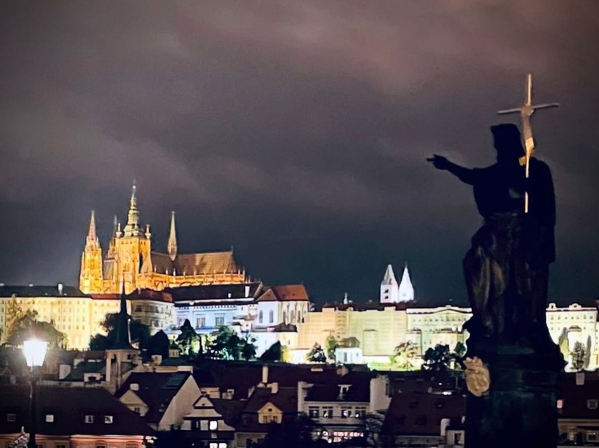 Nightscape of Prague, a silhouette statue pointing into the distance at a lot up building with spires