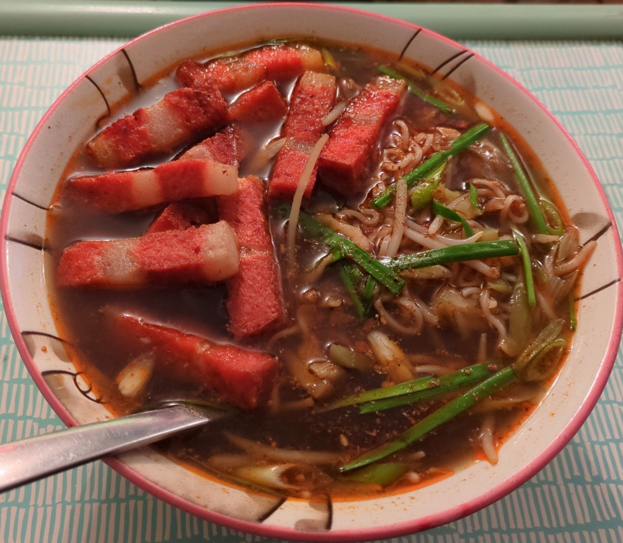 A bowl of hot and sour soup. The broth is a deep brown colour, made with stock, Chinkiang vinegar, Sichuan peppercorn, white pepper, soy sauce, and chili oil. Within it are ramen noodles, beansprouts, spring onions, garlic chives, and zha cai, which are all visible on the right side of the bowl; on the left are several pieces of Everbest vegetarian layered meat, air fried and then added as a topping before serving. There is also a further sprinkling of Sichuan peppercorn and a drizzle of sesame oil over the right side of the bowl; both can only be seen very faintly.