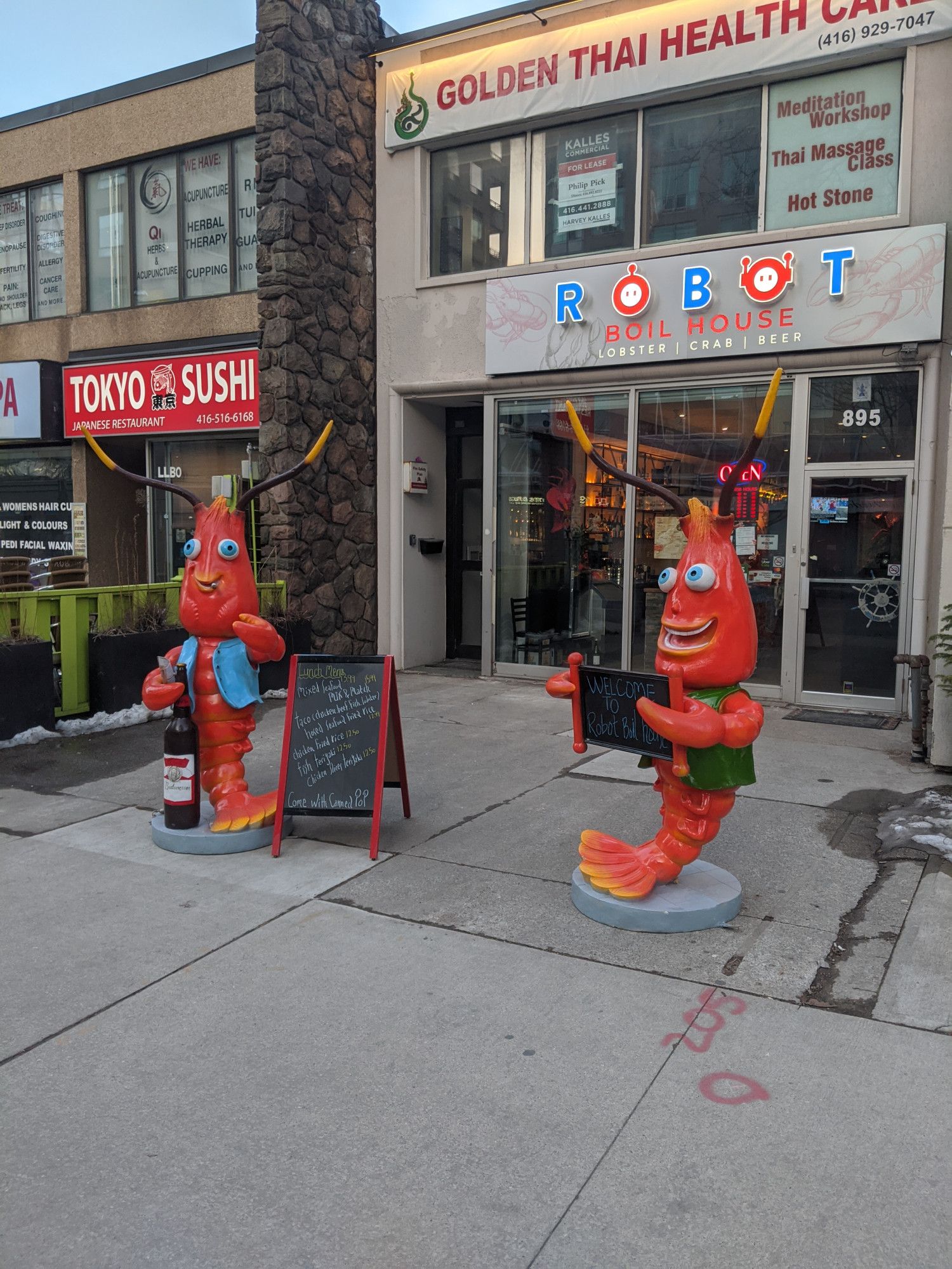 Two anthropomorphic crayfish statues sit outside a restaurant location in a small downtown plaza. The sign on the front reads "Robot Boil House: Lobster | Crab | Beer."