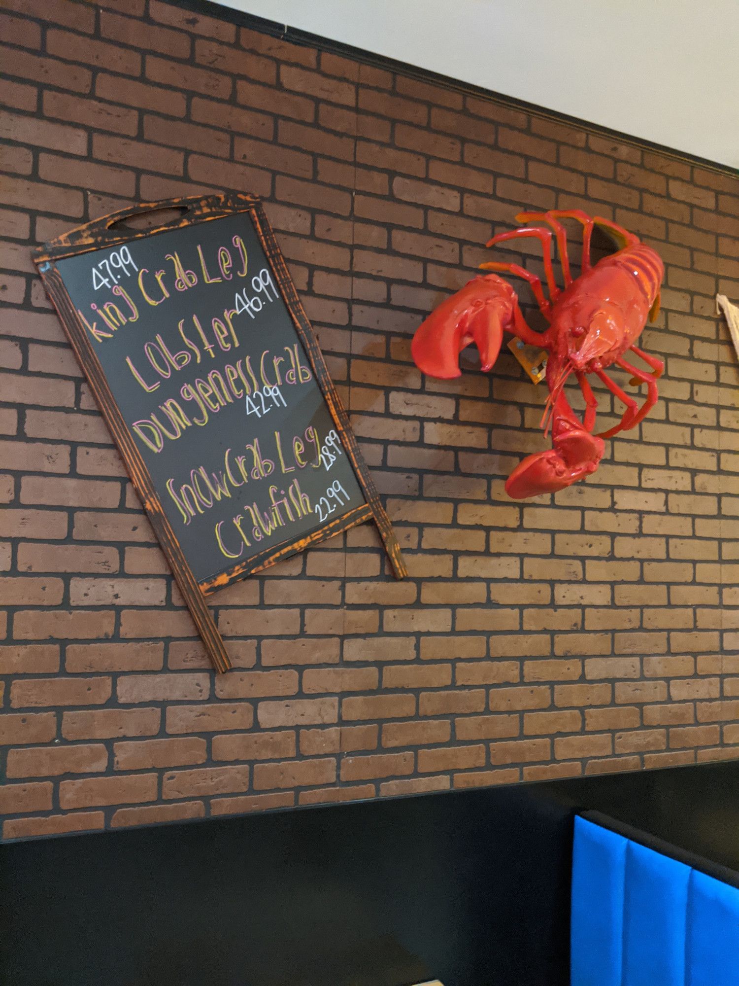 A wall of the Robot Boilhouse, covered in fake brown brick wallpaper. A large plastic lobster is affixed to the wall, next to a sandwich board that lists the day's seafood offerings (king crab leg, lobster, dungeness crab, snowcrab leg, and crawfish).