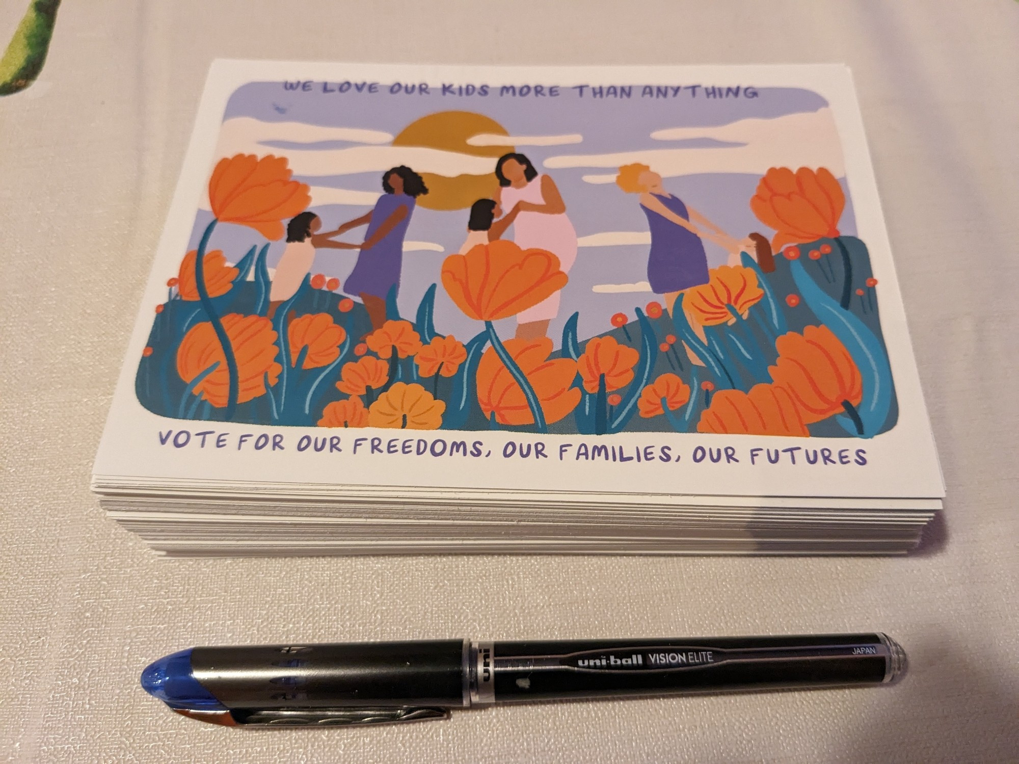 A stack of postcards with a pen. The postcards have a drawing of people and orange flowers and read "We love our kids more than anything. Vote for our freedoms, our families, our futures."