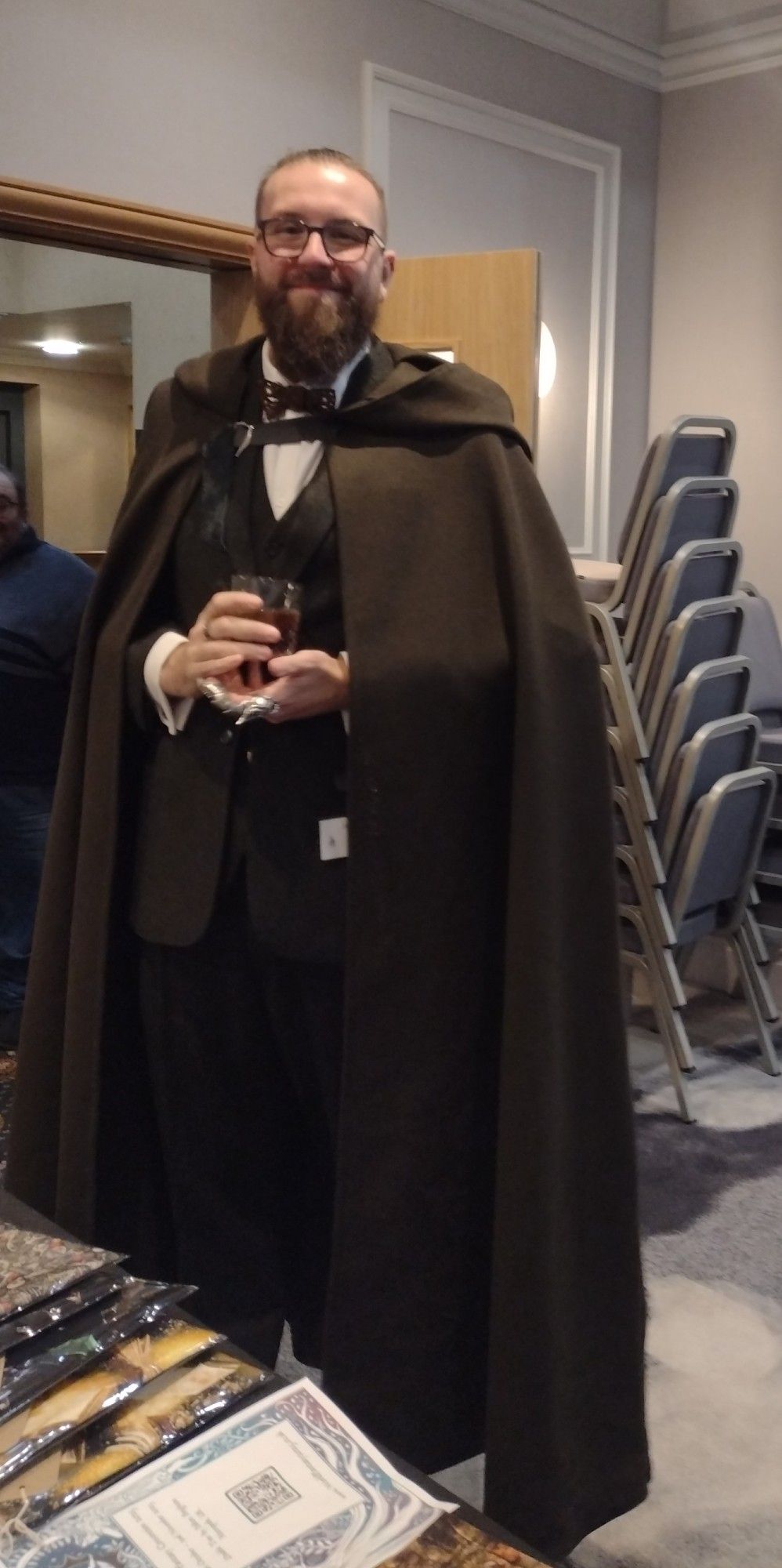 Dan Hanly, a tall person in a simply stunning cape over a suit with finger furniture. As he holds a pint, his claw ring cradles it. He will definitely solve your mysteries.