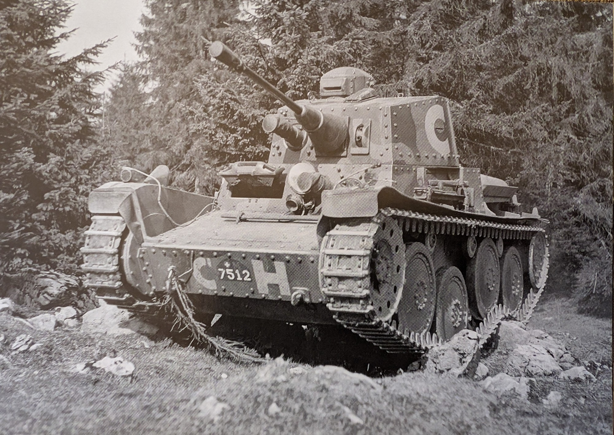 A picture of a Panzerwagen 39, a light tank used by the Swiss Army. It's on uneven, grassy land, with some rocks protruding through. The tank itself is perched on some of these rocks, causing the tracks to rise up halfway along the vehicle.

In the background just behind the tank is a pine forest.