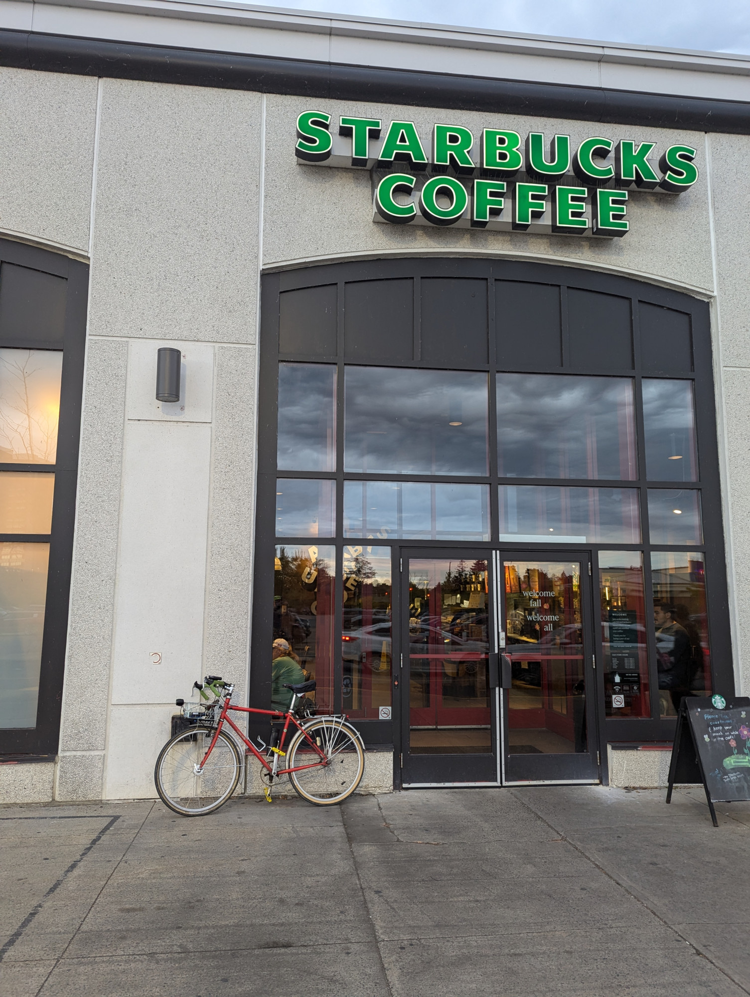 A red bike with a front basket, viewed side on, leaning against the front of a Starbucks. The coffee shop doors are to the right of the bike