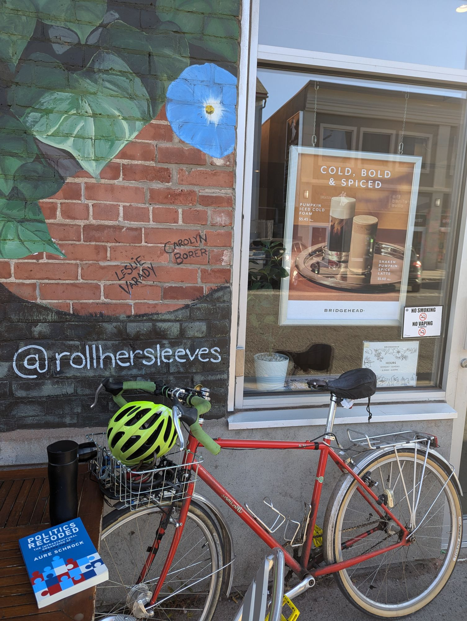 A red bike with a front basket, a helmet in the basket, leaning against a coffee shop wall and window. A table in the foreground with a travel mug and a book on it. Book title is Politics Recoded.