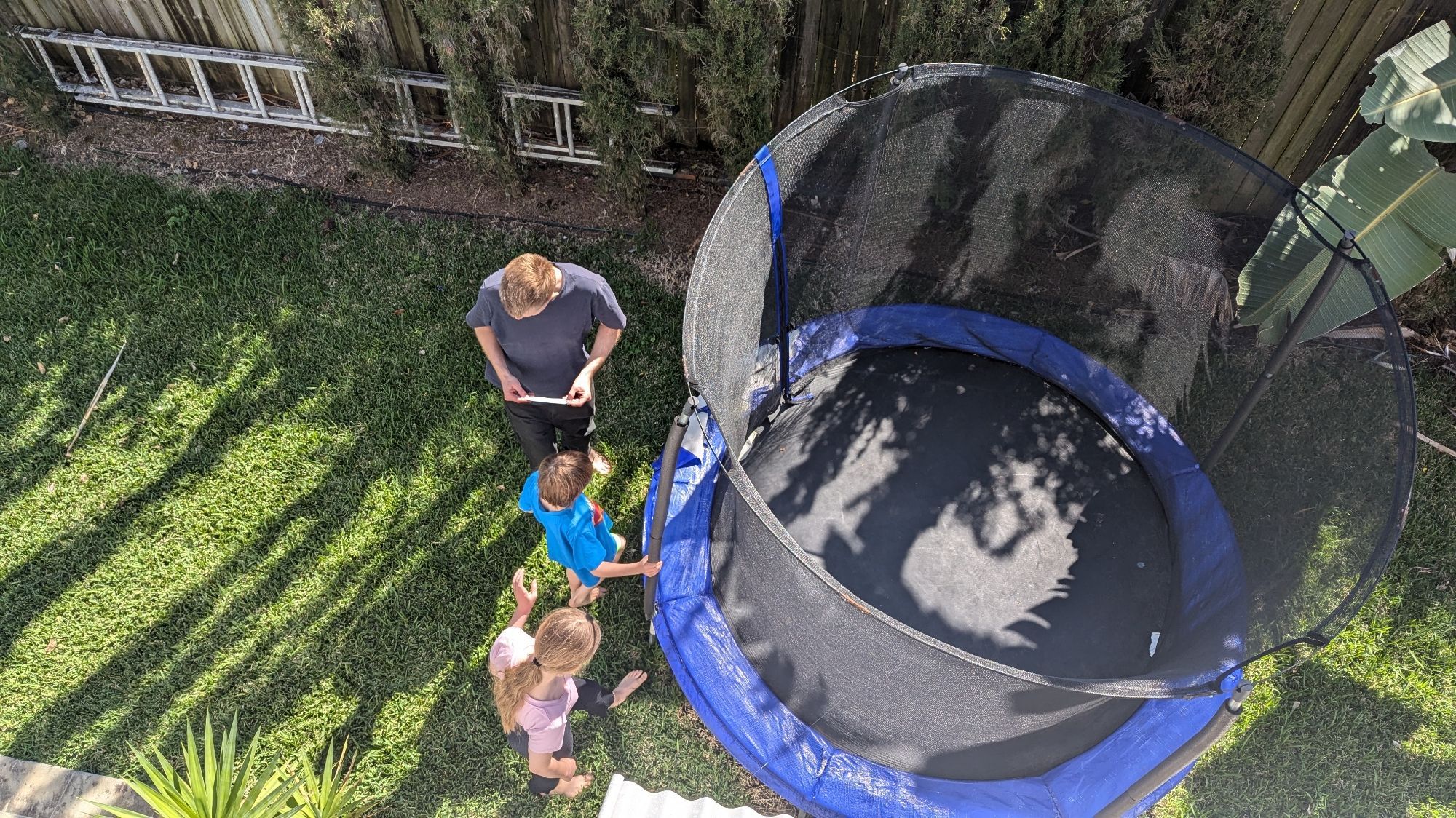 Man and two kids out in the sun next to a trampoline, looking at a small piece of paper