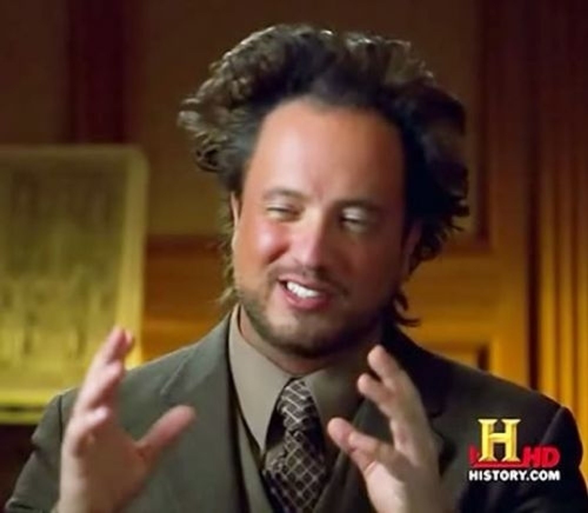 "Aliens" meme guy.  Dressed in a gray suit with taupe button up and tie.  A white man with receding hairline that is fluffed wildly at the sides and back has his hands raised with palms facing each other as he says "Aliens"

The background of the room he is seated in has wood paneling.