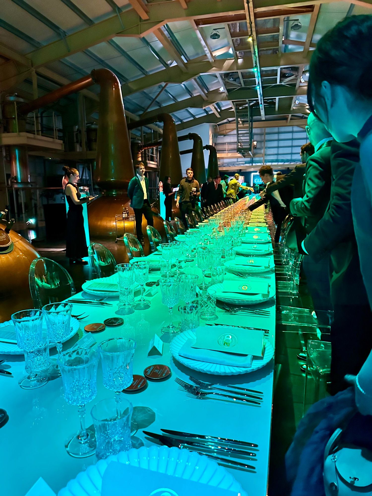 An extremely long table set for a formal dinner, running between the copper pot stills in The Glenlivet distillery. People are dressed up in their glad rags. There are coloured lights shining onto the table to up the mood: gold, blue, and a teal green (The Glenlivet's brand colour). The copper stills are gleaming. There are menus on the plates embossed with a gold '200' design, because this dinner is to celebrate the distillery's 200th anniversary. It's all very fancy indeed.