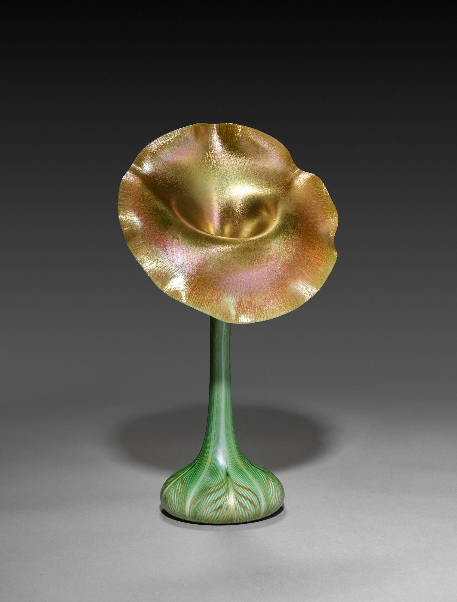 This is a photo of a blown glass vase shaped like a jack-in-the-pulpit.  The base of the vase looks like the bottom of a squashed teardrop and has gold stylized leaf pattern on light emerald colored glass.  The neck is the same green and is very narrow.  The top is an open circular flower significantly wider than the base.  The rear of the flower is lifted up, and the front is angled down.  The open flower is metallic in appearance and is in shades of gold, peach, orange, and pink.