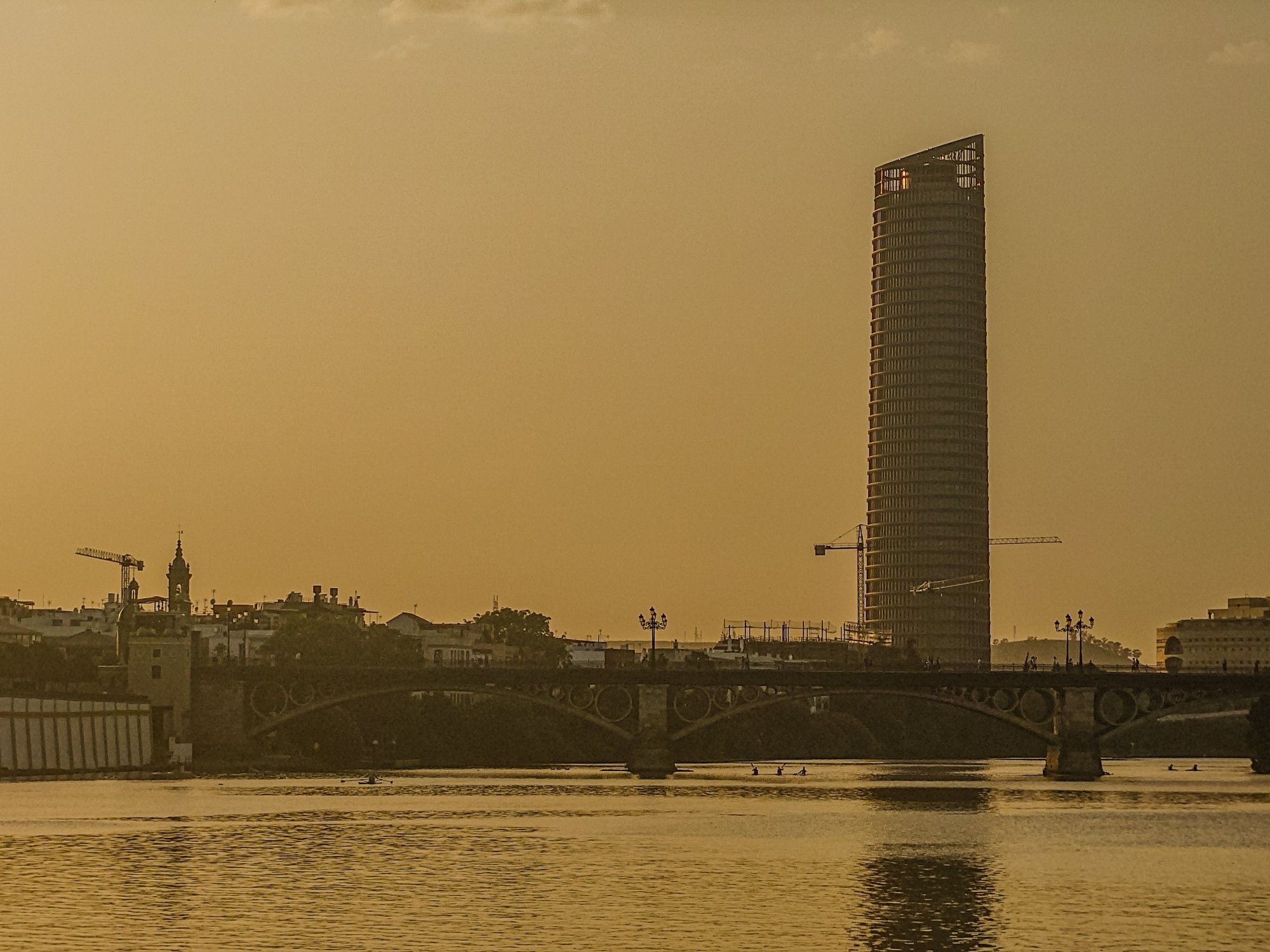 A photograph of Seville taken in the dusk. The image looks across a river towards a bridge and a large building behind it. There is a lovely golden light in the air and the building is reflected in the river.