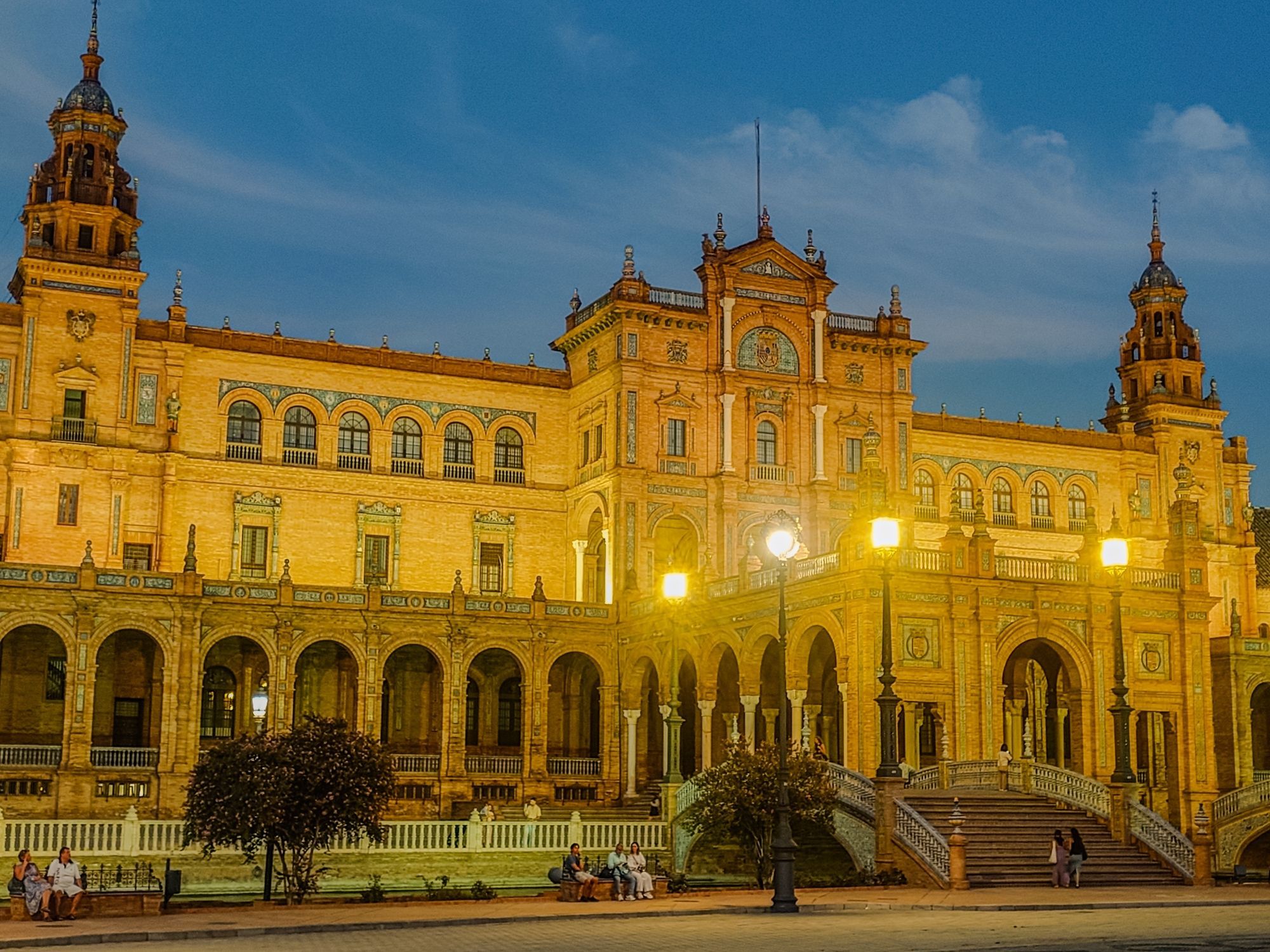 A photograph of the Plaza de Españia in Seville. The photograph is taken in the dusk with the lights on. It is a very grand building that was built for an exhibition in the 1920's, and is now part of a large public park. It was used in the first Star Wars prequel movie as the scenes on Naboo.