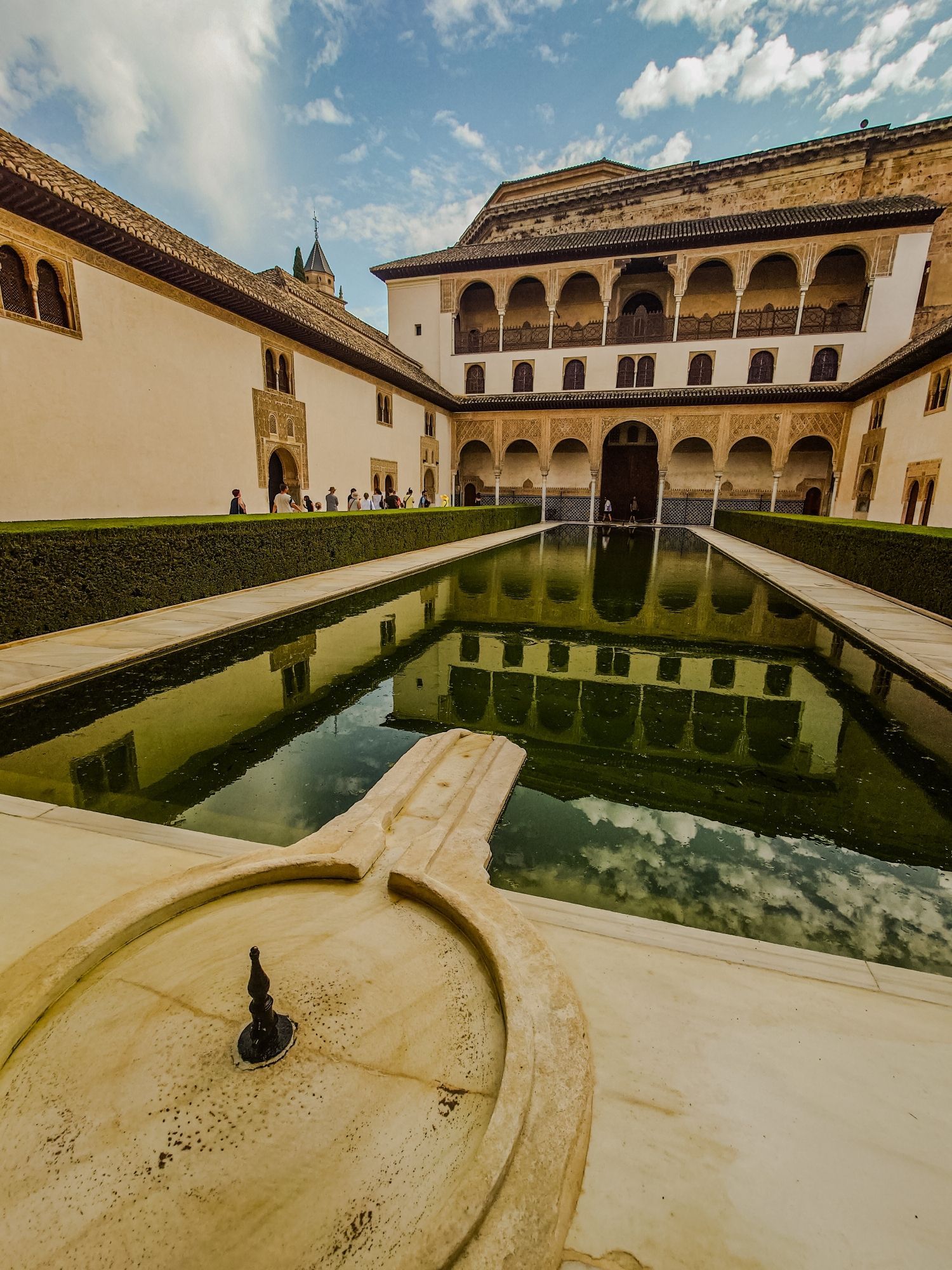 A photograph of a courtyard in the Alhambra. The courtyard has a large pool in it, and a building supported by columns next to it. The archways are engraved with intricate patterns and dates back to the days of the Caliphate in Spain. As it's Granada in the summer, it is a hot and clear day.