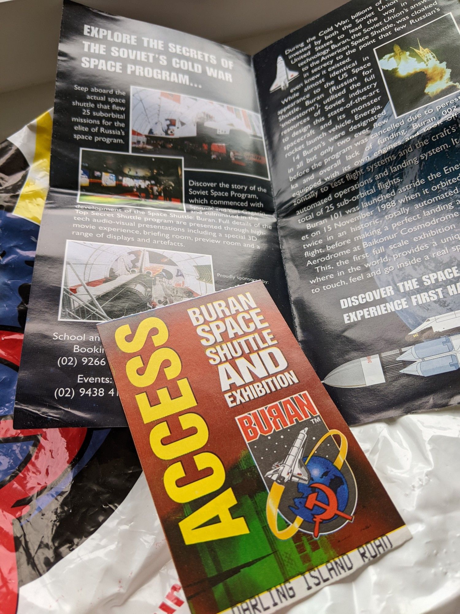 A pile of souvenir stuff - a plastic gift shop bag, an informational leaflet, and the entry ticket to see the Buran Soviet Space Shuttle. The latter has the printed piece 'Darling Island Road' showing that this exhibition was in Sydney, Australia.