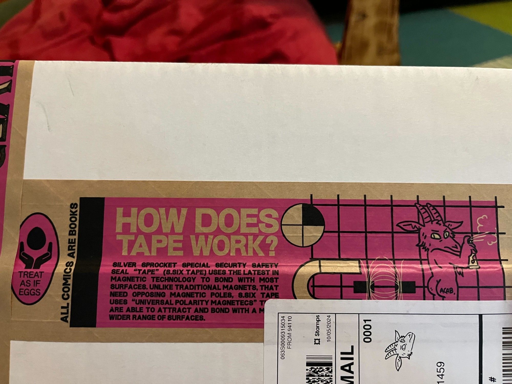 A strip of brown packing tape on a package with printing:
An icon showing two hands holding an egg, with the text "Treat as if eggs"
Text: "All comics are books"
A large block of text:
How does tape work?
Silver sprocket special security safety seal "Tape" (s.Six tape) uses the latest in magnetic technology to bond with most surfaces, unlike traditional magnets, that need opposing magnetic poles, s.Six tape uses "Universal polarity magnetecs" that are able to attract and bond with a much wider range of surfaces.
A diagram showing a magnet, two rectangles with magnetic field lines, and a goat (the Silver Sprocket mascot) spraypainting a wall, with its back turned to show its (human) butt with "ACAB" on the right cheek